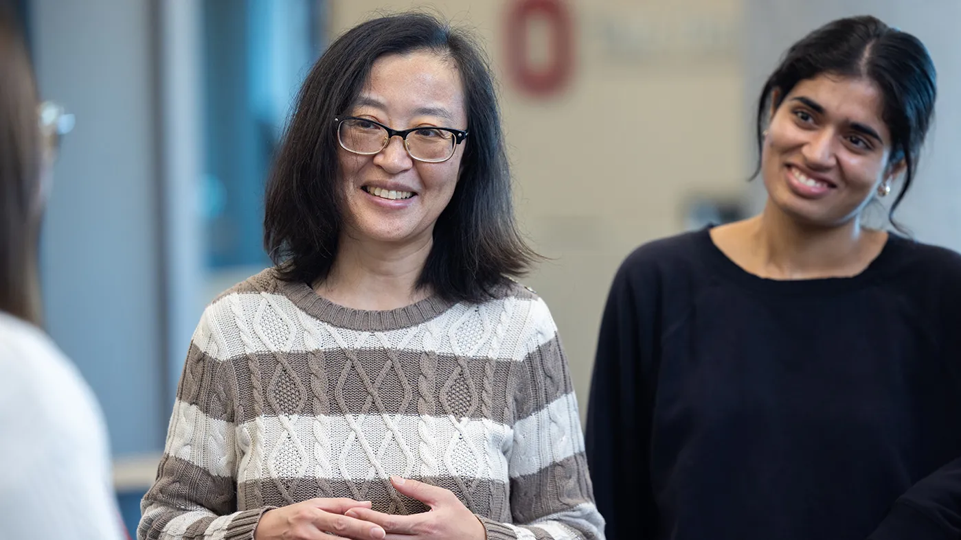 A woman of Chinese descent and slightly behind her, a woman of Indian descent, smile as they listen to an off-screen colleague speak