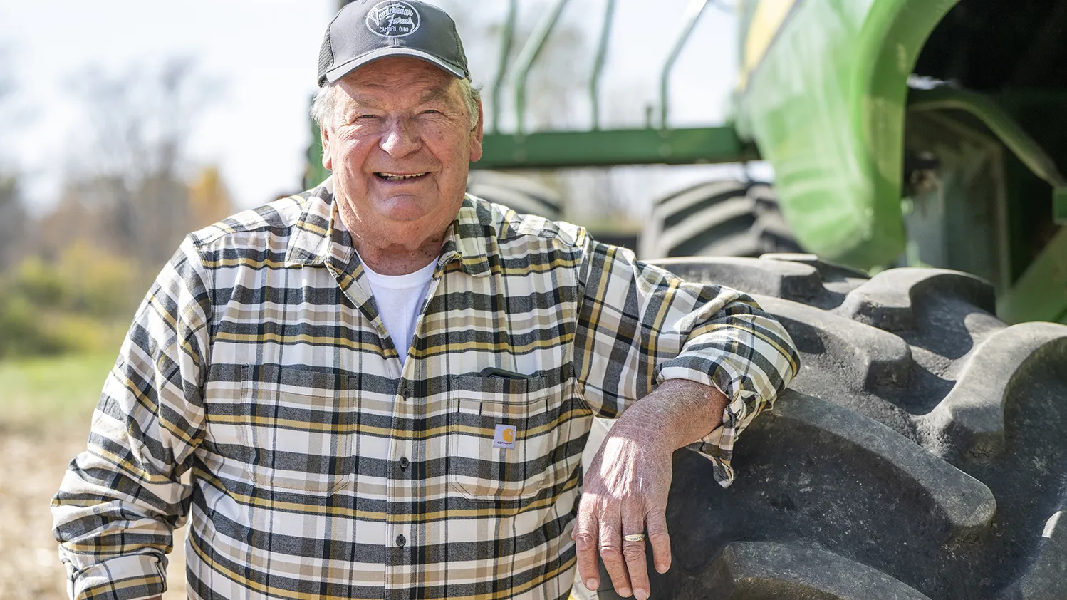 Len Vonderhaar is an older white man wearing a plaid shirt and smiling happily as he leans on a wheel of his tractor