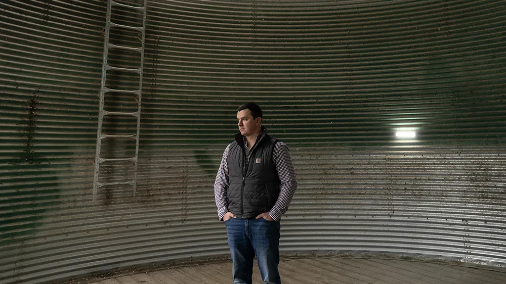 Will Hamman, a young white man wearing jeans and an insulated vest over his plaid button-down, stands inside an empty grain silo on his family farm in Ohio. The structure is corrugated metal maybe five times as wide as he is tall, with a peaked roof. A ladder and big flood light are too his right.