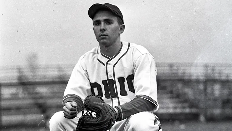 In a historical photo, a baseball player crouches and wears a serious expression as he is photographed. The front of his jersey says Ohio and his mitt is thick with an extra large thumb portion.