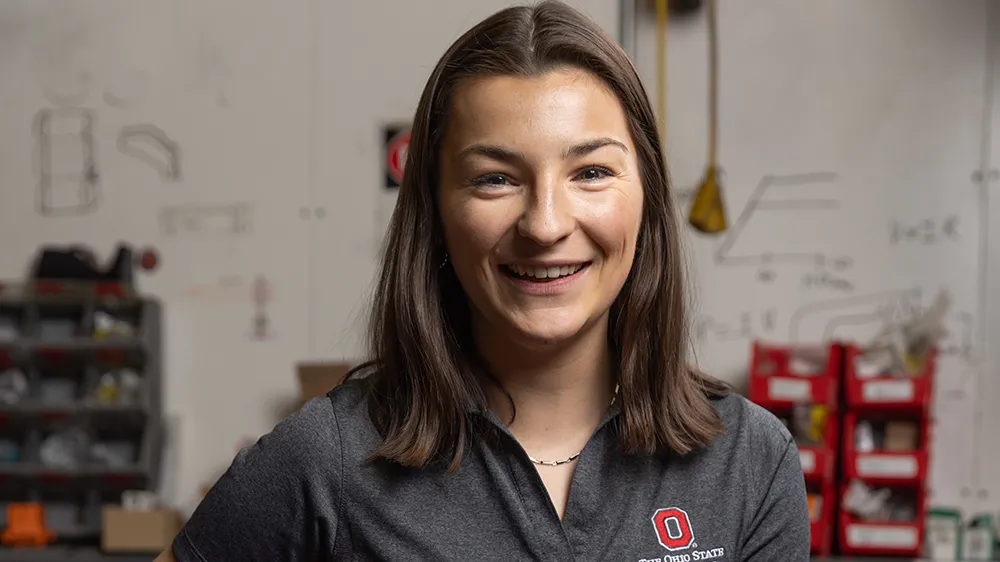 Laura Friedmann wears an Ohio State polo shirt branded Center for Automotive Research, as she smiles in this portrait in a motorcycle laboratory. She is a young woman with shoulder-length hair and her smile says she is genuinely thrilled to be photographed.