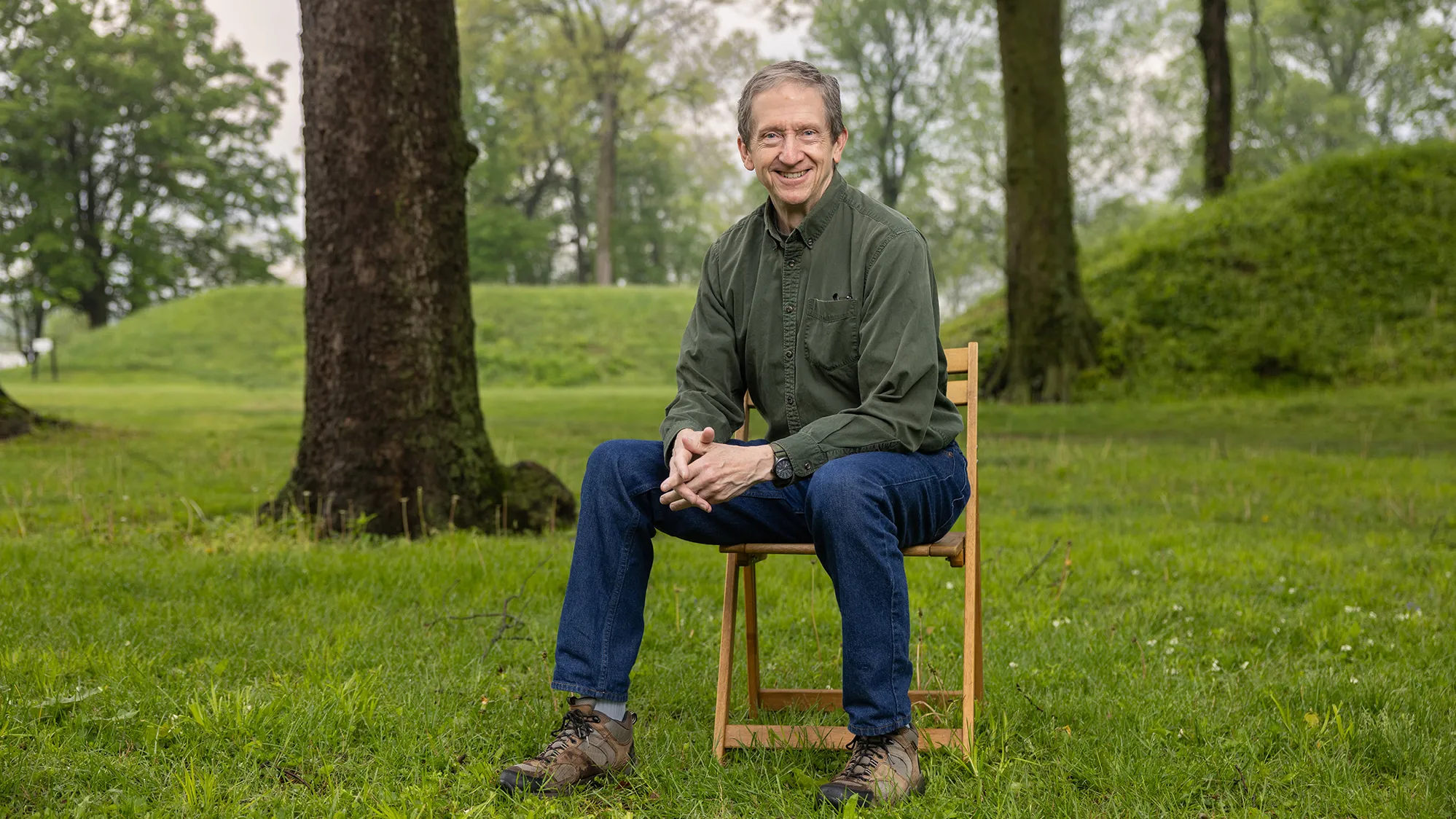 Bradley Lepper, a gray-haired white man in jeans and an earthy colored button down, sits on a chair at the Great Circle Earthworks. Around him are trees and behind him, grass-covered mounds rise. The portions of the mounds in the photo look to be about as tall as an adult, or at least shoulder height.