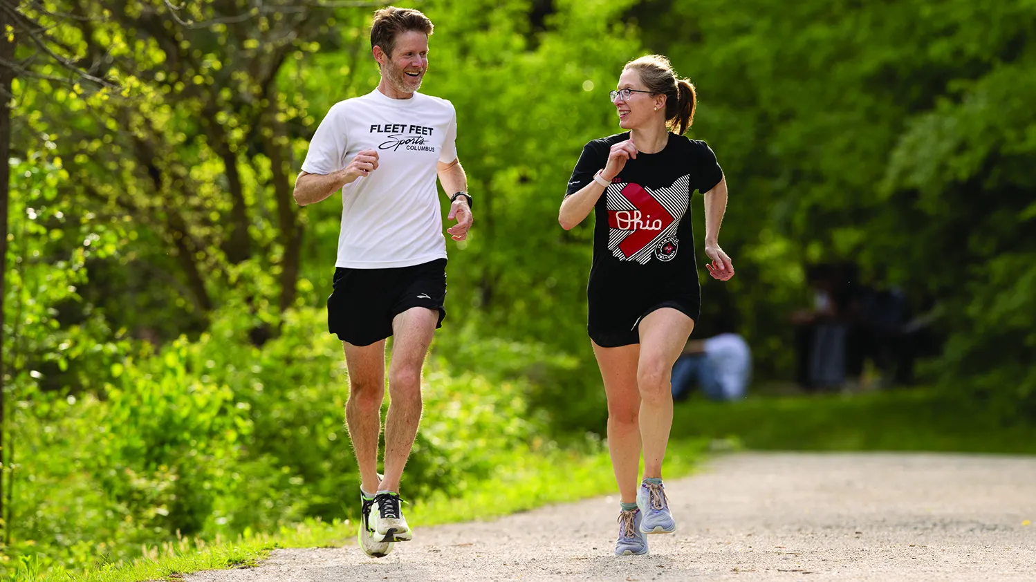 A woman and man talk as they run down a bicycle trail surrounded by trees and lush vegetation.
