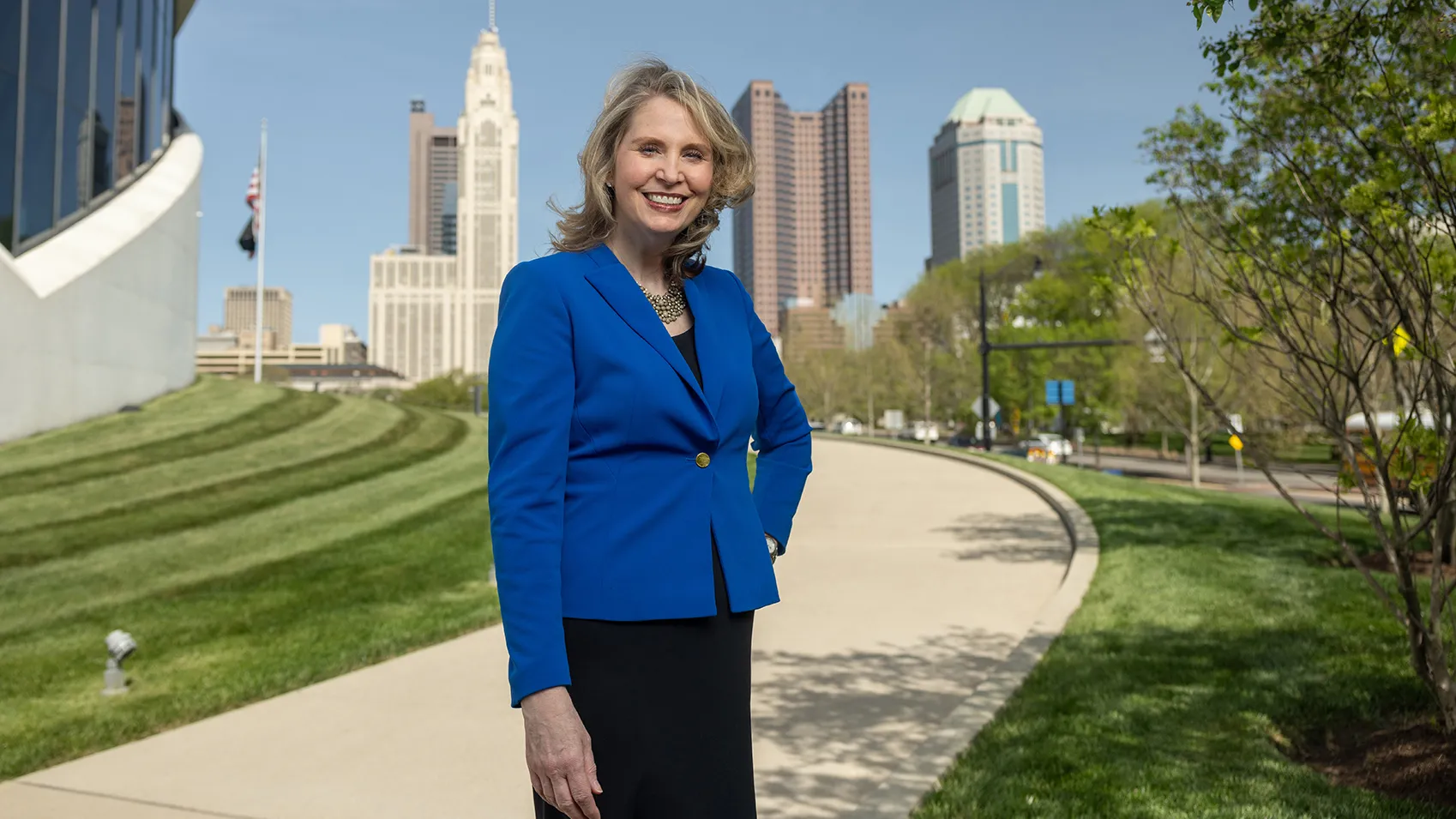 Amy Edwards Taylor smiles and stands with one hand on her hip as she poses for this photo. Behind her is the edge of the Veterans Memorial Museum and farther away, downtown Columbus. She has shoulder length blond hair, wears a business suit and smiles like she's genuinely happy to see you.