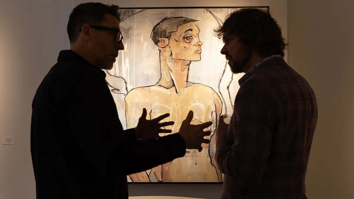 Two gallery guests are silhouetted in front of a modern painting of a man. The painting is lit up behind them.