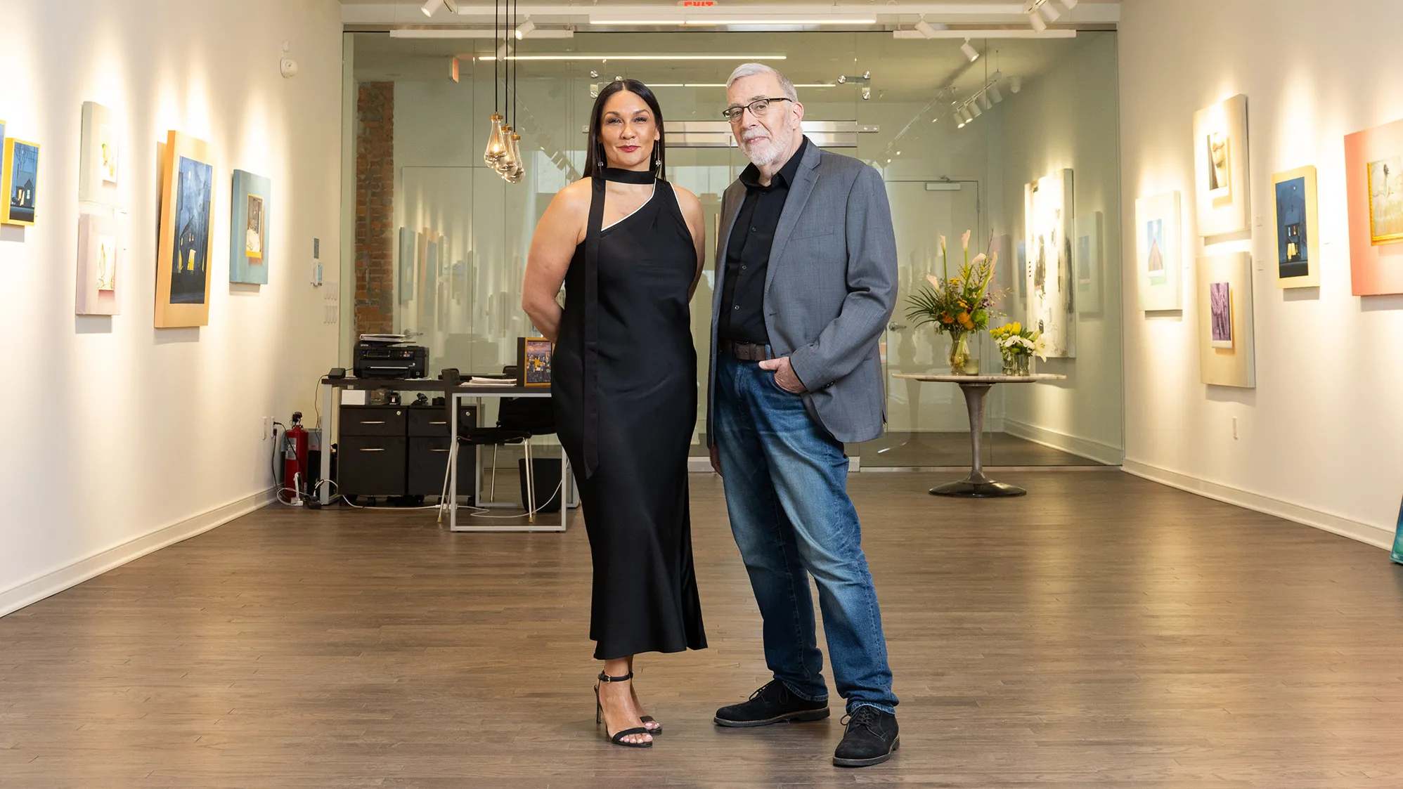 Michelle Brandt wears a black dress and Duff Lindsay wears jeans and a sports coat in a new art gallery. They seem elegant and thoughtful as they pose standing next to each other and slightly smiling.