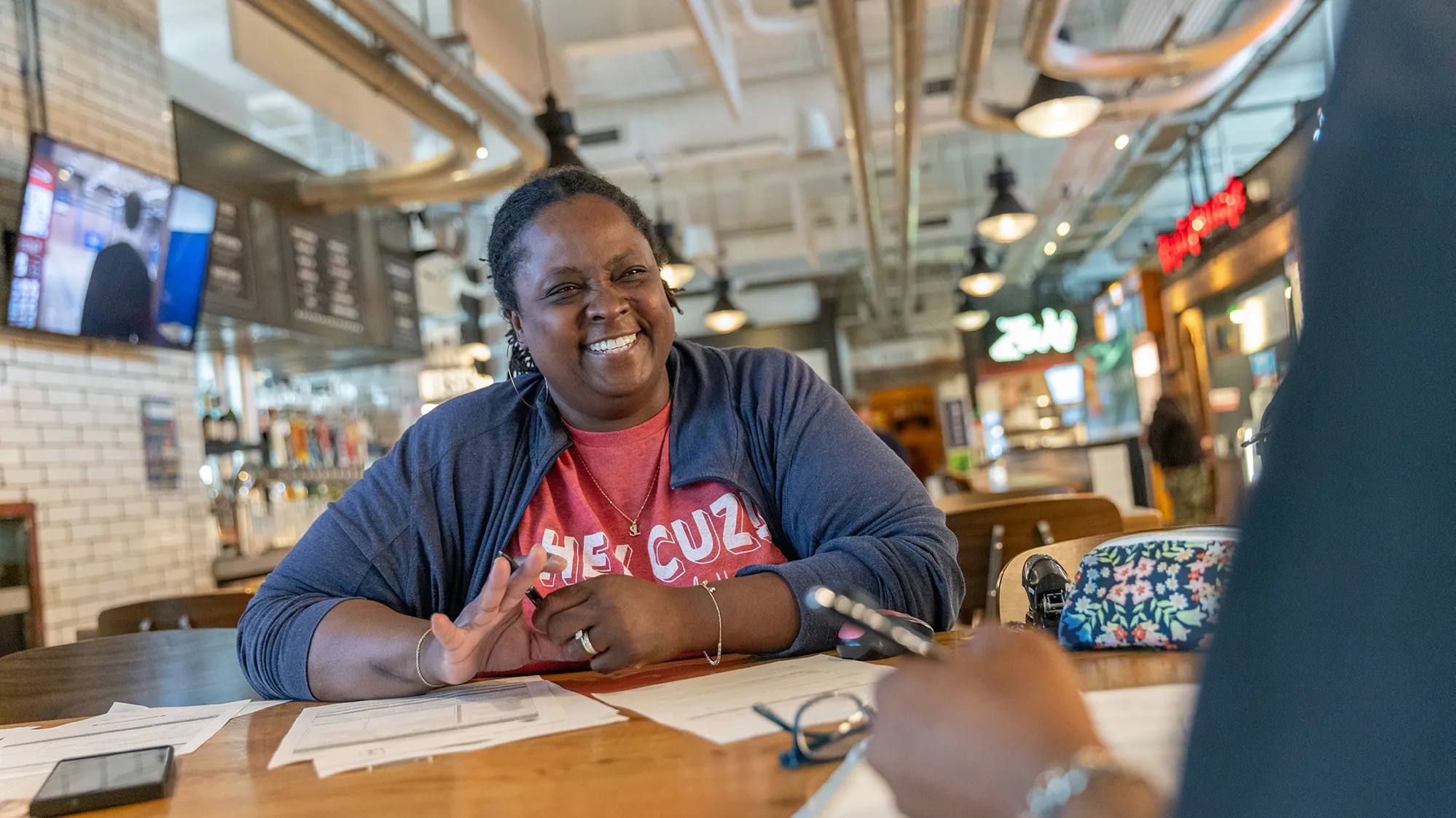 Sadaya "Daisy" Lewis sits at a hightop table to onboard an employee. This food hall is where her popular Modern Southern Table restaurant is located. There is exposed, shiny ductwork, white brick walls and a clean, modern feel.
