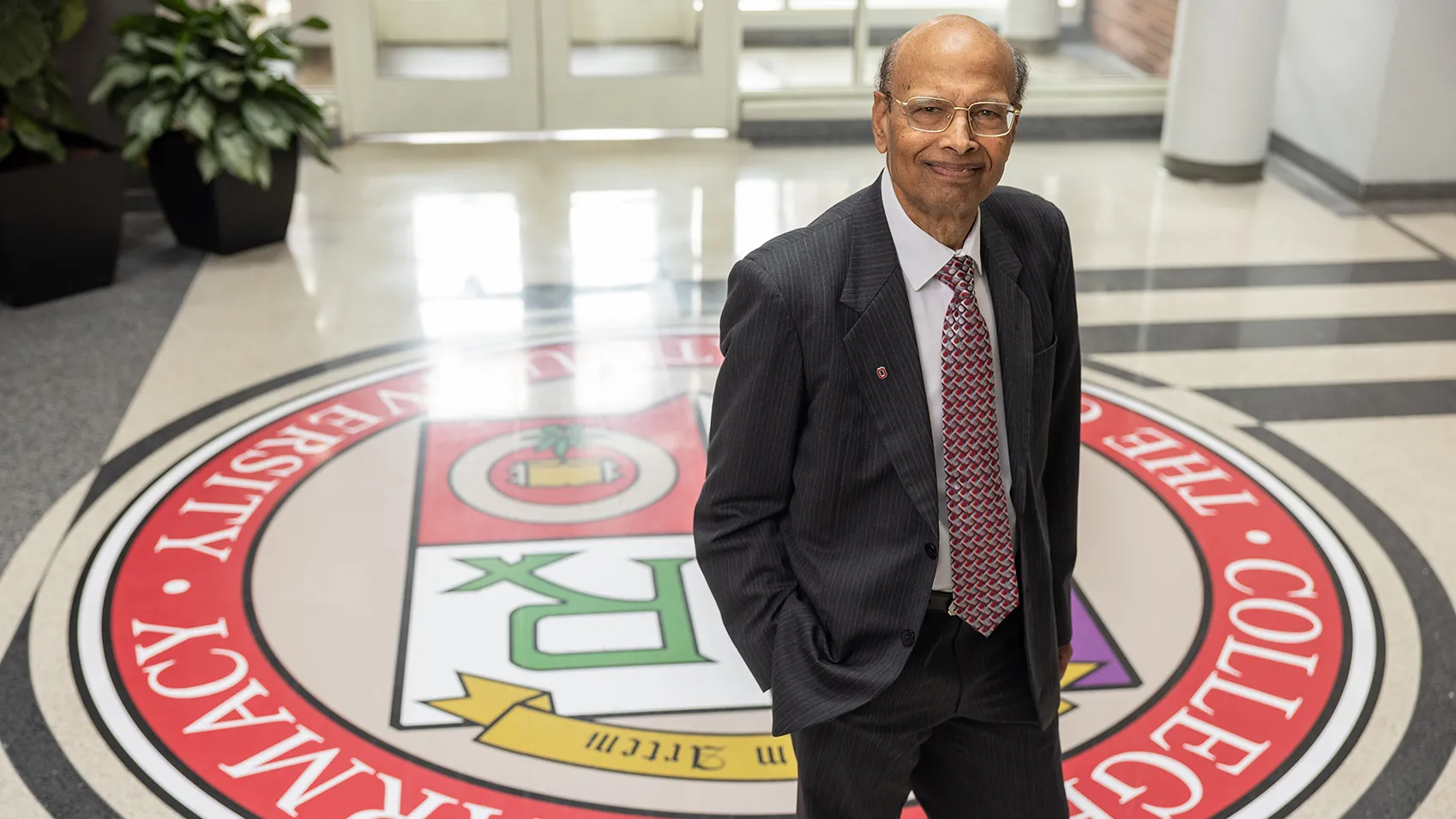 Milap Nahata, an Indian man, poses for a portrait in the lobby of the pharmacy college at Ohio State. Its colorful insignia is painted on the floor, adding an interesting background. Nahata looks calm, confident and friendly.