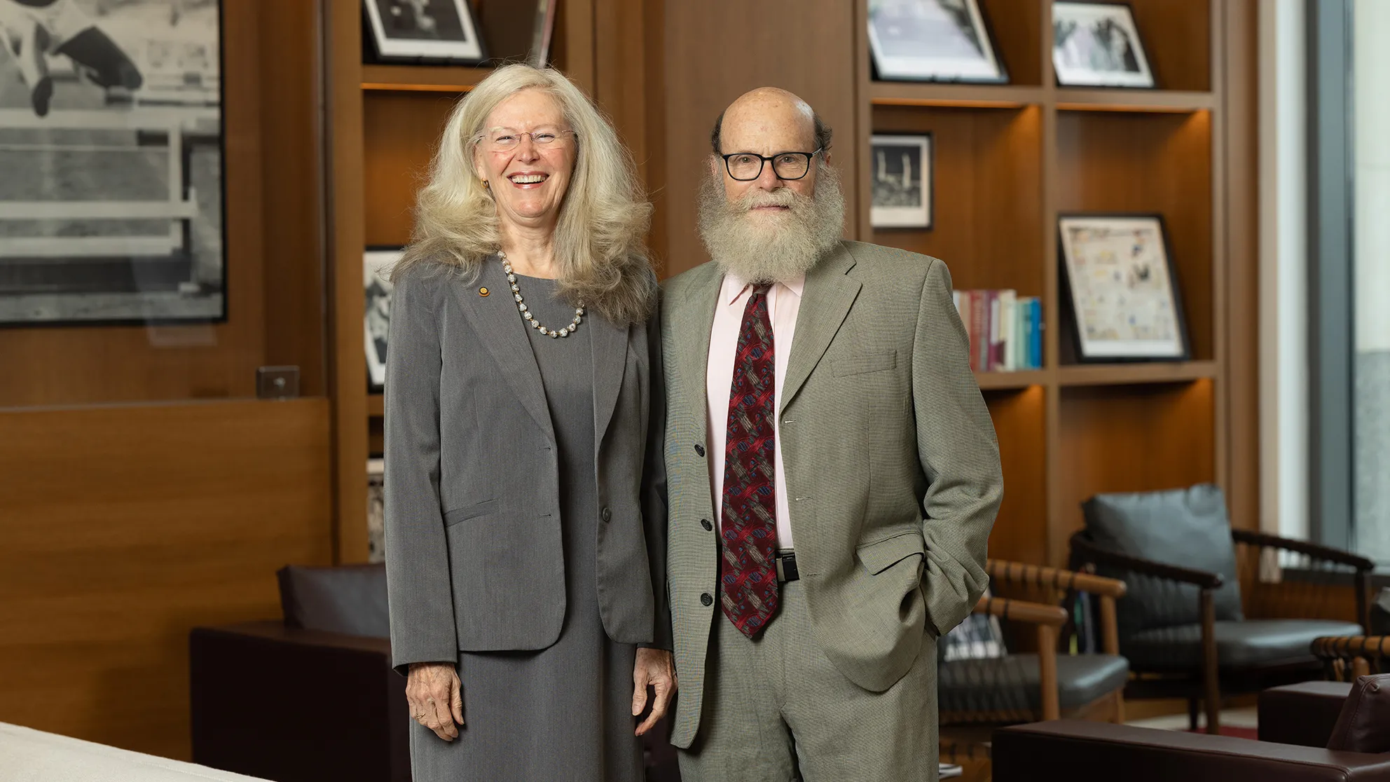 Gay Miller and Joseph Rosenblatt are a white couple who are both dressed in suits. She has long, pretty hair and is laughing. He has a lush beard and glasses. They are in the lobby of a university building. Around them are Ohio State memorabilia on wooden shelves and chairs.