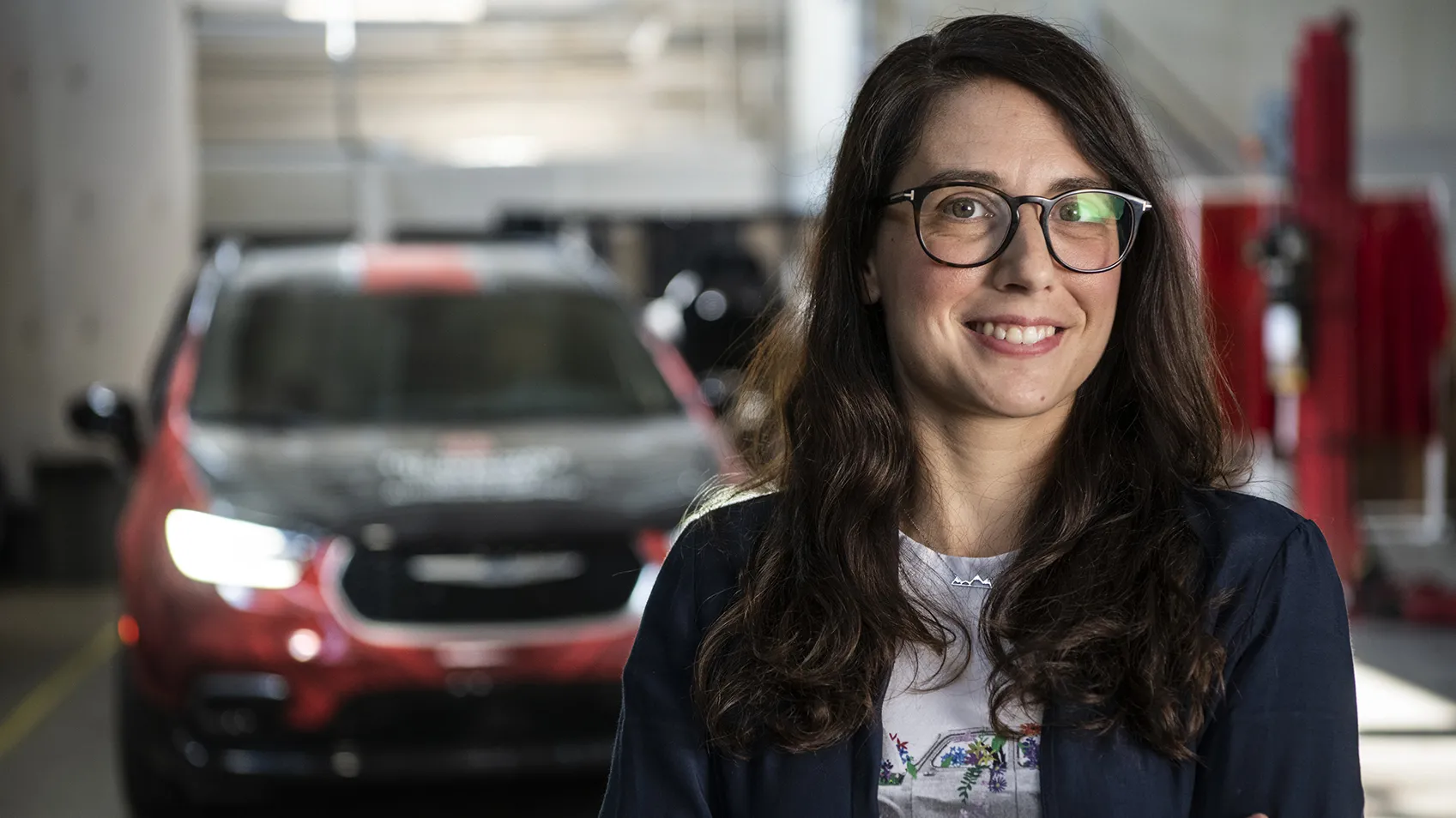 Stephanie Stockar, a white woman with glasses and long brown hair, smiles for a portrait in the automobile laboratory where she works, she wears T-shirt and sweater. Behind her is an electric car.