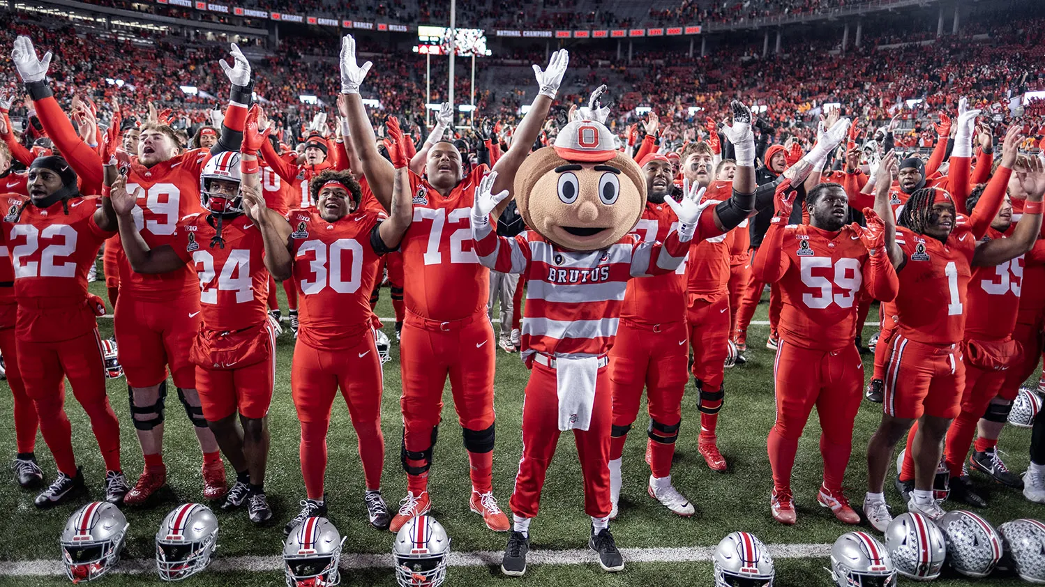 Brutus stands slightly in front of the football team, all on the field in Ohio Stadium, with crowded stands far in the distance. They're all raising their arms and smiling as they sing Carmen Ohio.