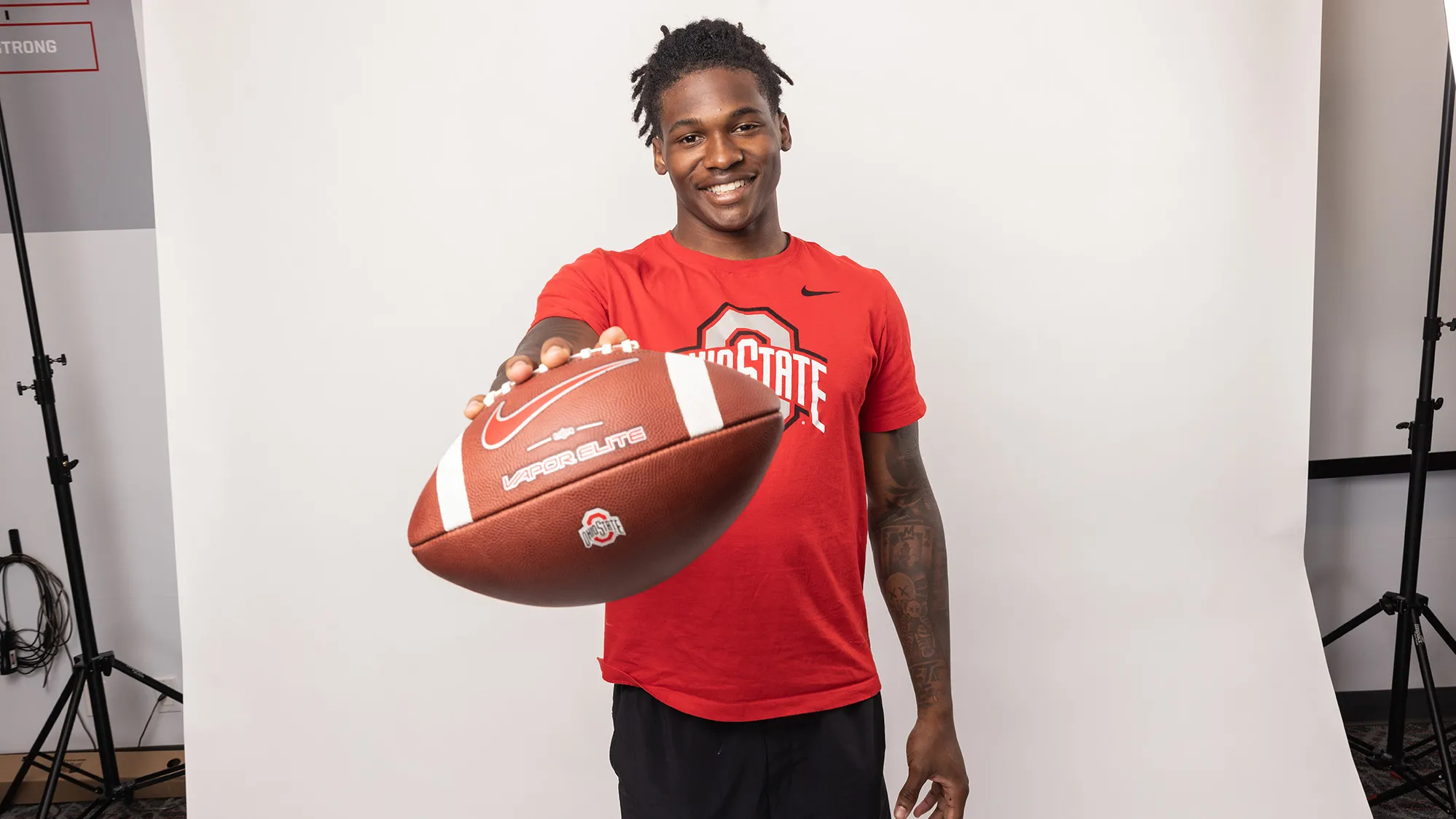 Ohio State football player Jeremiah Smith poses for a photo holding out a football. He's an attractive young black man, smiling with his hair pulled up in a ponytail and tattoos on his arms.