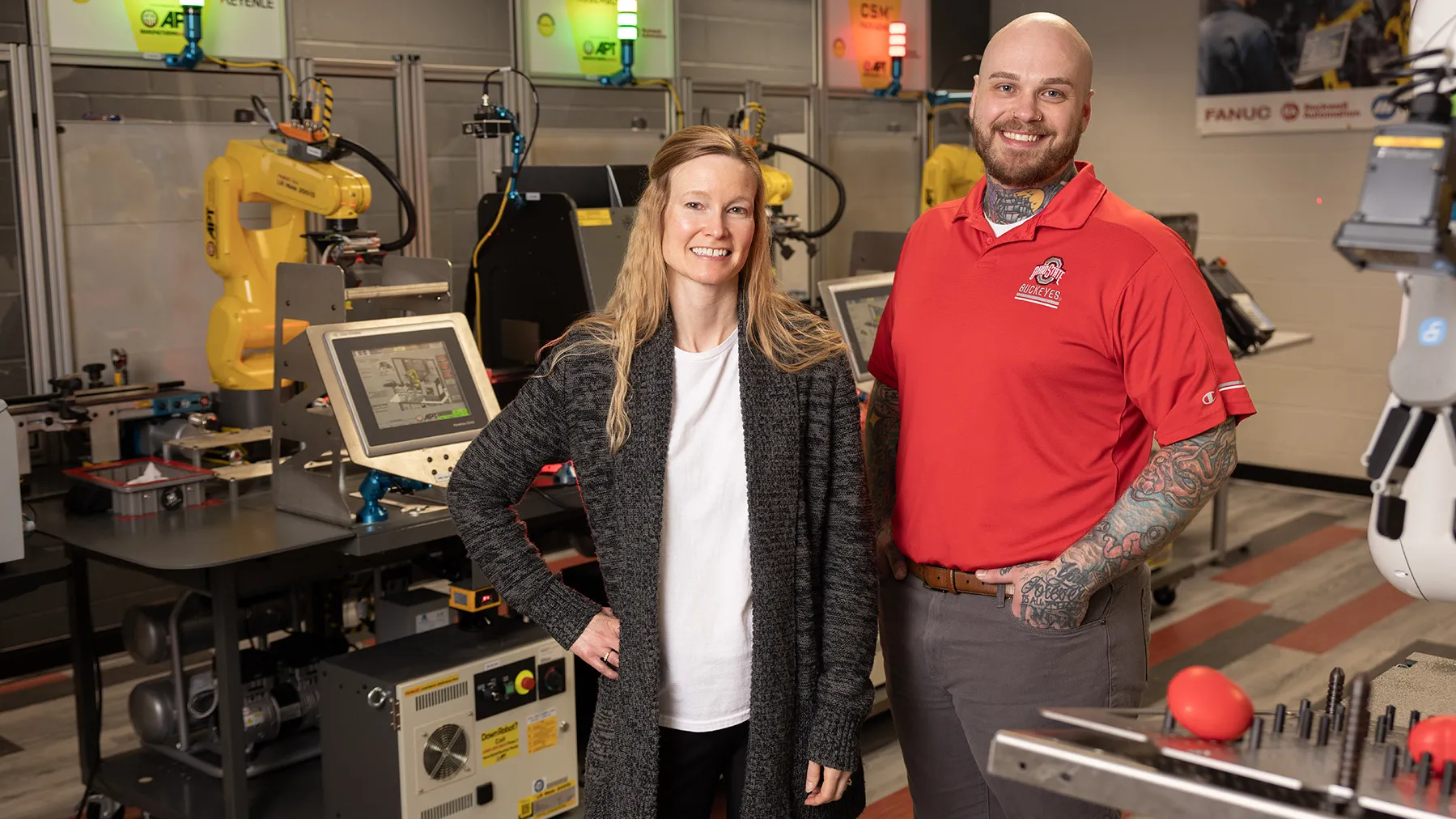 In a hands-on classroom at Ohio State with computers and automated manufacturing equipment, two people pose for a photo. They both seem happy and at home in the lab. The woman, who is white and half a foot shorter than her former student, has long blond hair and stands with one hand on her hip. The man is Zack Ernest, and he stands with a hand tucked in his hip pocket.