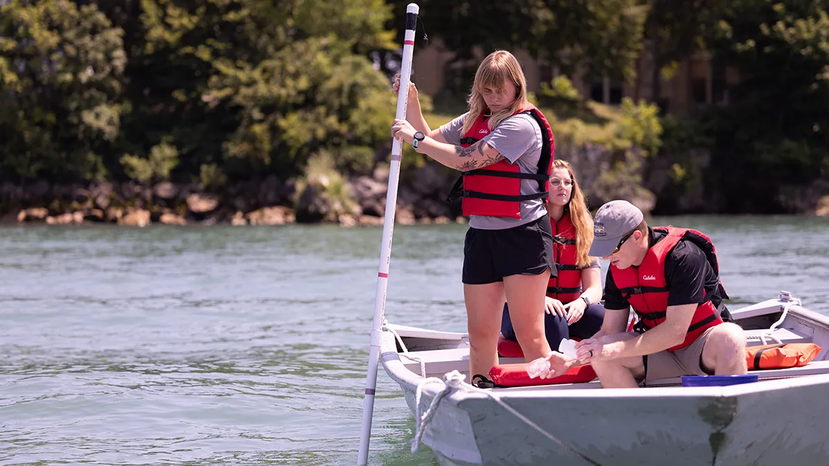 In a canoe on Lake Erie, a young blonde woman stands and reaches a pole into the lake. A man takes notes while sitting. A third person is behind them but is barely seen.