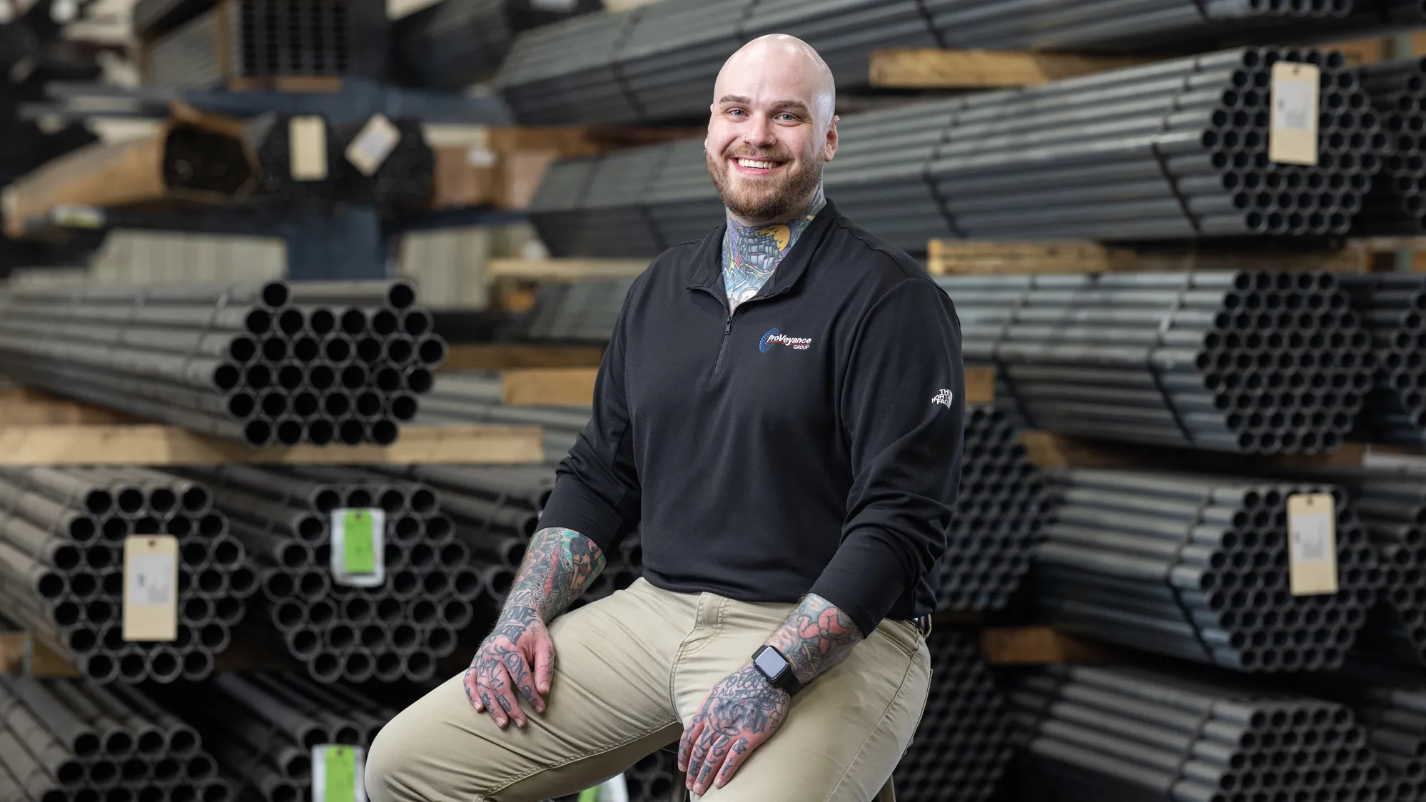 Zachary Ernest, a tall white man with shaved head, neat beard and colorful tattoos on his neck and forearms, smiles as he sits on a stool on the factory floor of Ashland Conveyor Products. He is in front of bundles of about 50 metal pipes each, stacked on shelving. 