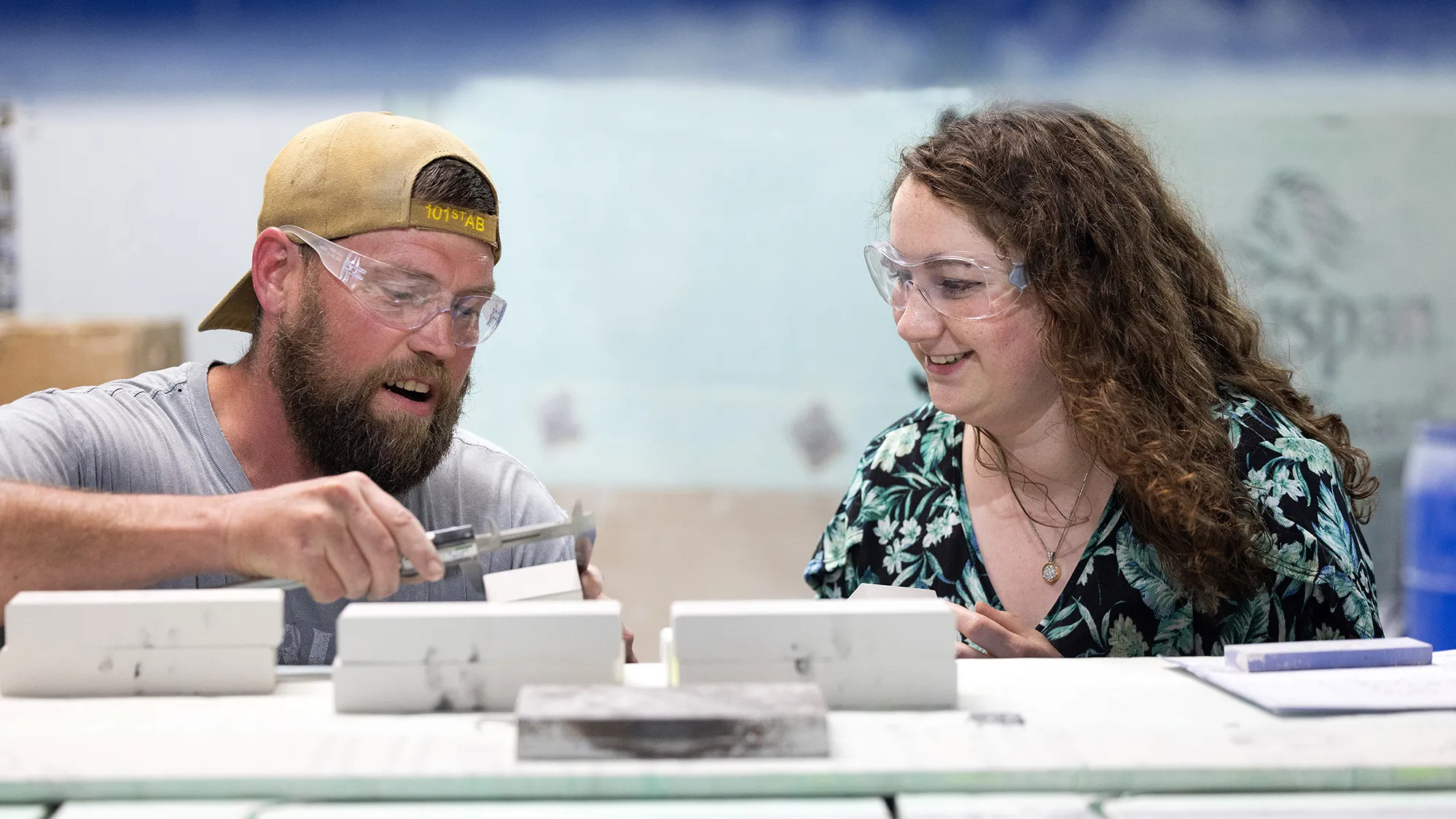 A man talks to the woman next to him. Both are white, wear safety goggles and are smiling. In front of them are blocks of ceramic. Not the ceramic like you use in dishes but less polished, industrial ceramics.