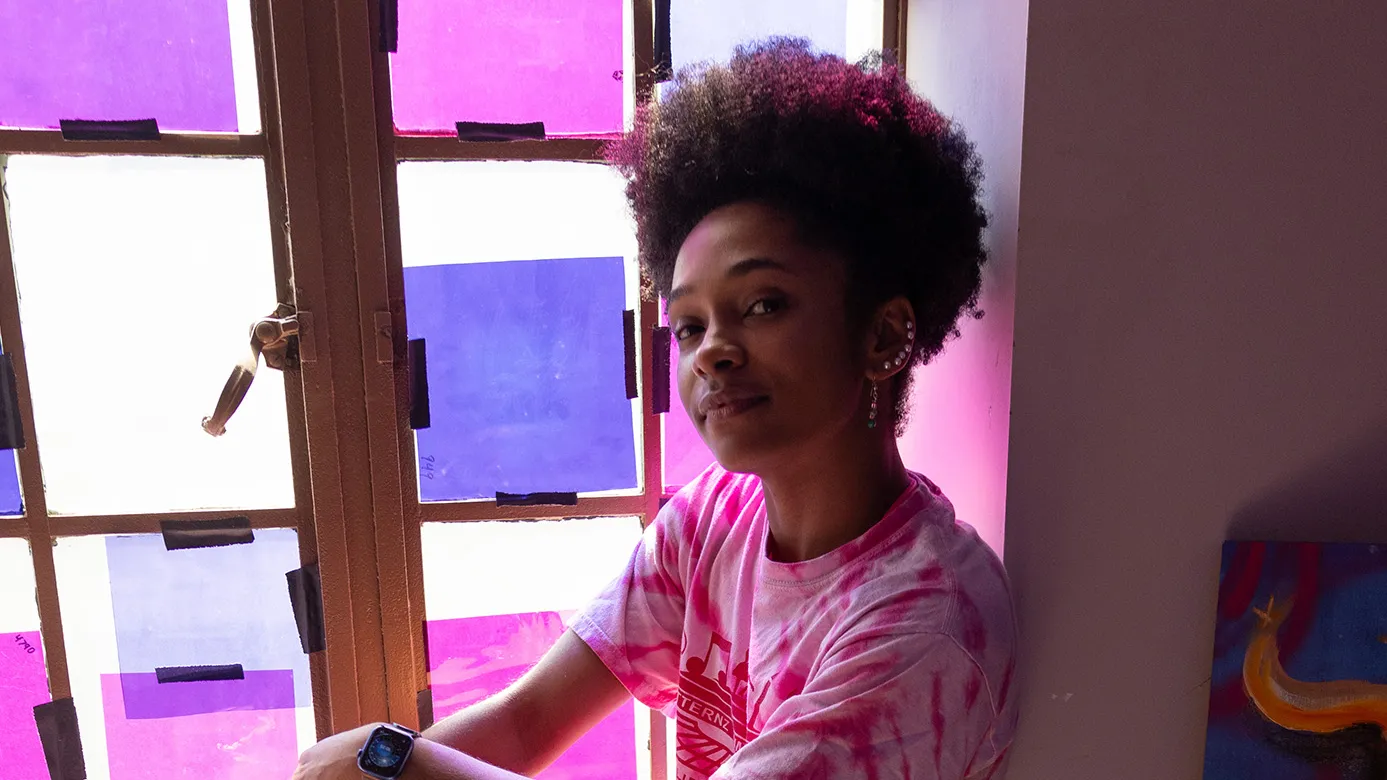 Brianna Rhodes, a pretty, young black woman, sits on a window ledge inside a dim room. Colored paper taped over the window shines colored light onto her. She's smiling with her eyes and wearing casual clothes that she can dance in. Her hair is tight curls on top of her head and she has a line of earrings up the ear the photo shows.