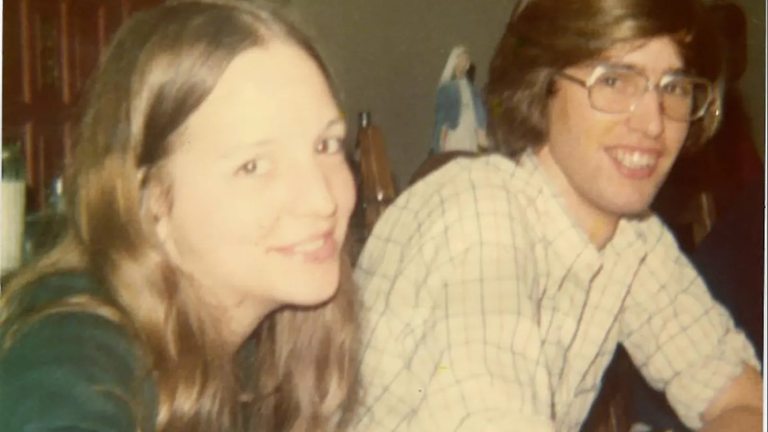 In this 1970s photo, a young white couple smiles at the person taking the picture, almost as if they're saying "if you really have to take this photo, go ahead." He wears glasses and she has long hair--their hair color almost matches.