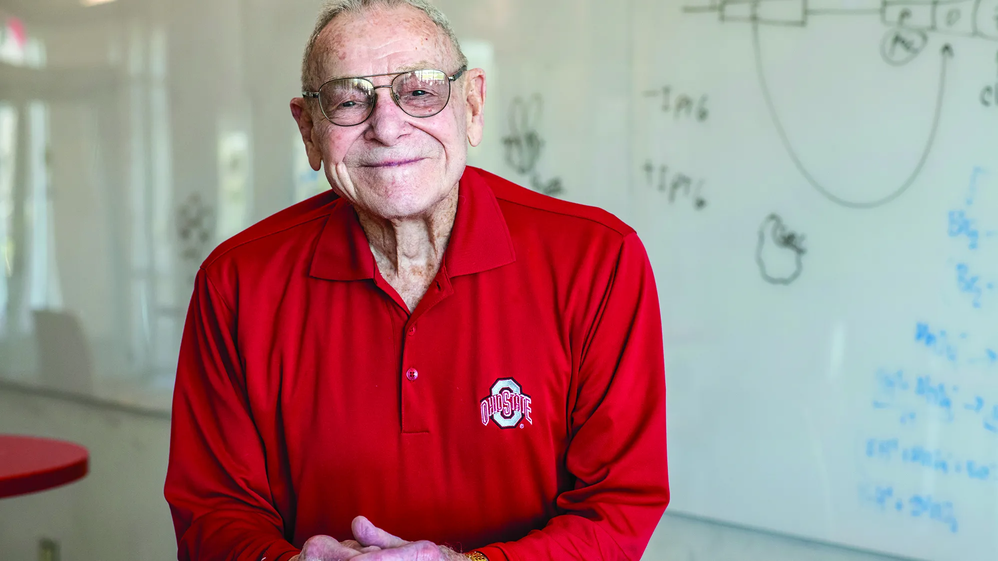 Merle Lashey smiles with closed lips, but not less enthusiasm than a big grin, in a classroom on the Marion campus. He is an older white man wearing a scarlet Ohio State golf shirt, and behind him is a white board with hand-drawn diagrams.