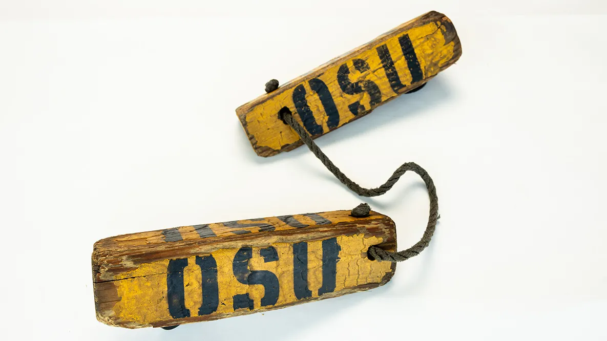 Two weathered, long triangles are tied together by a rope. They have the letters OSU stenciled on them and are shaped like this so that they can be placed on either side of an airplane wheel to stop it from rolling.