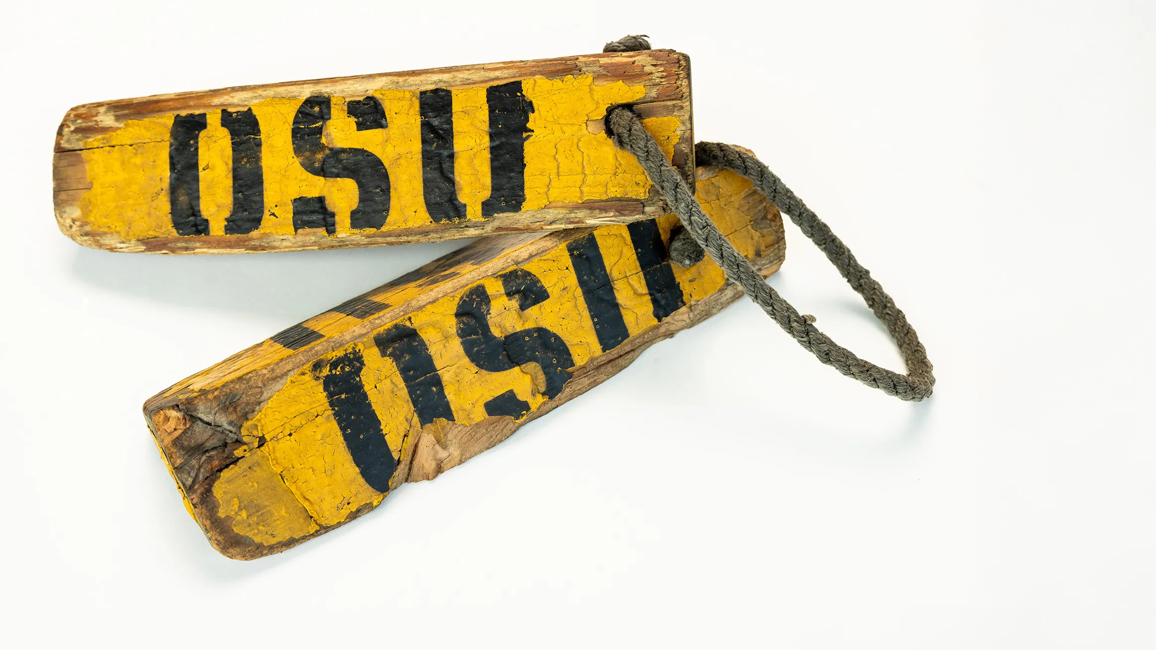 Two weathered, long triangles are tied together by a rope. They have the letters OSU stenciled on them and are shaped like this so that they can be placed on either side of an airplane wheel to stop it from rolling.