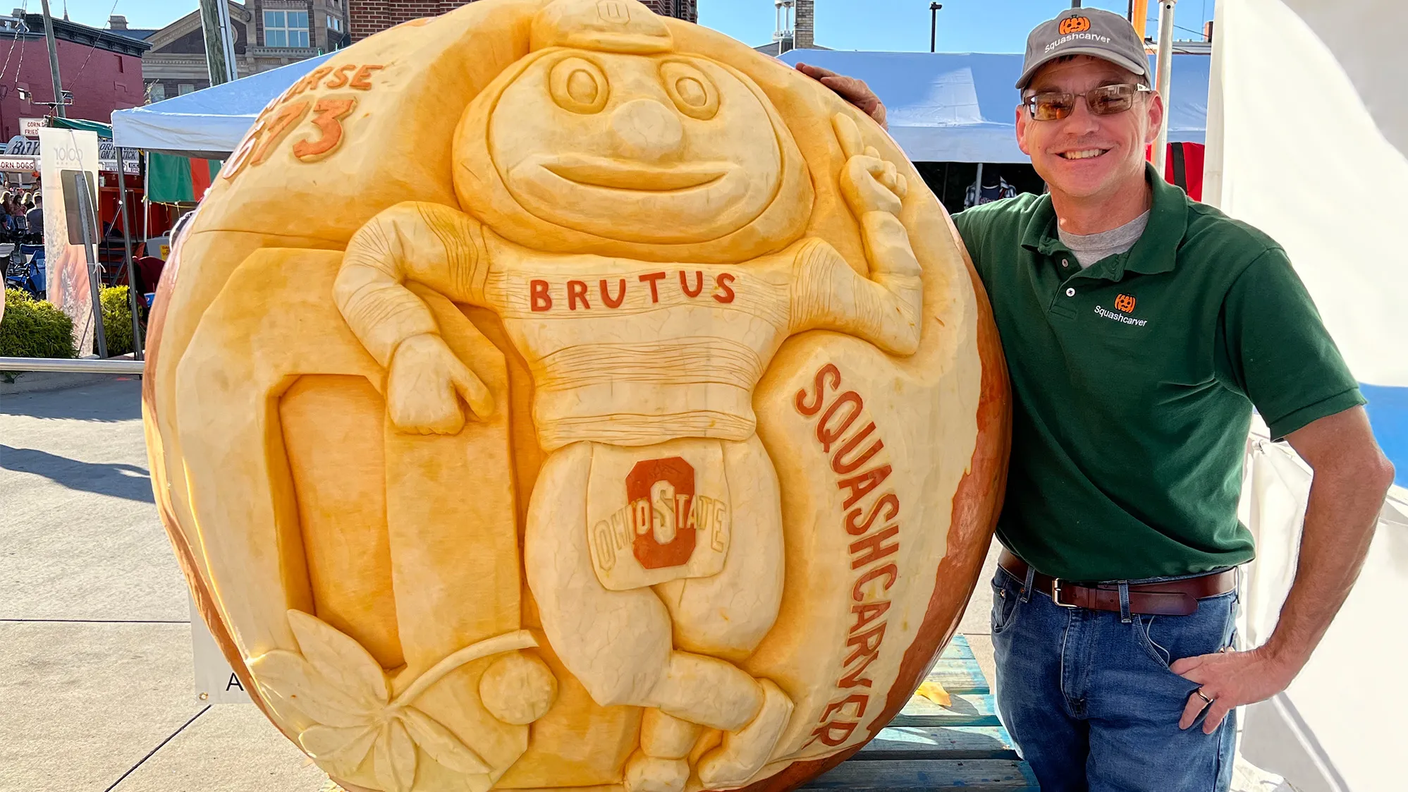 Gus Smithhisler grins as he shows off a giant pumpkin he just carved. It has Brutus Buckeye leaning on a Block O. Gus is a white man wearing sunglasses and a ballcap in this photo he shared.