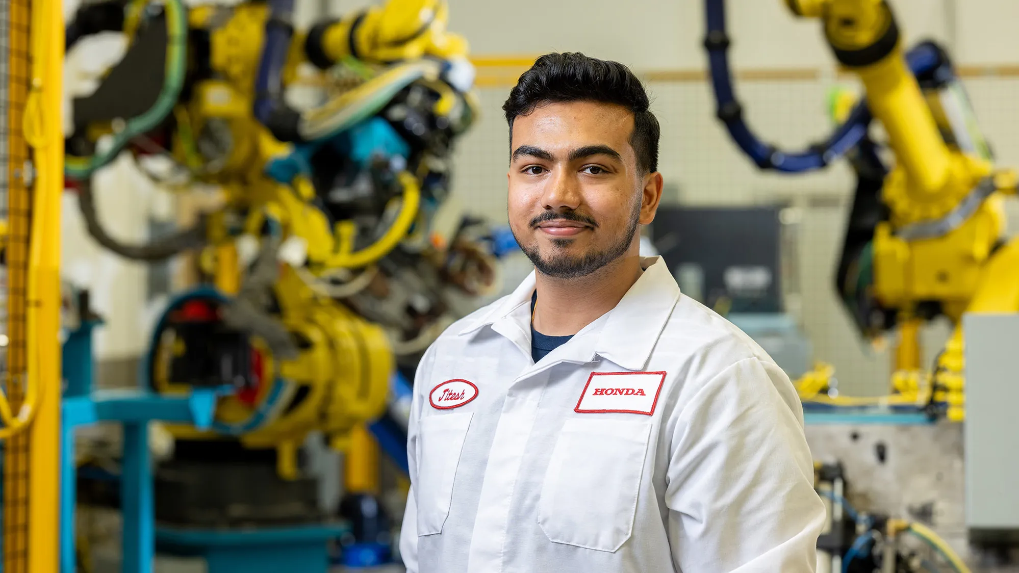 Jitesh Vidhani poses for a photo wearing his Honda uniform and in a hightech factory. Yellow, heavy duty robotic arms and other machinery is working in the background. Jitesh is slightly smiling, with his eyes and mouth.