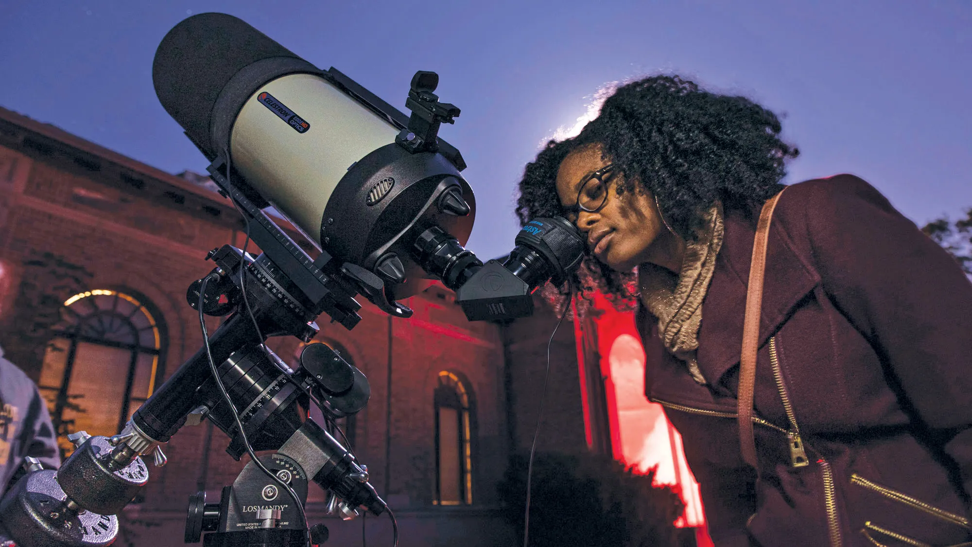 Caprice Phillips, a young black woman wearing her hair curly, a fall coat and glasses, peers into the scope of a telescope. Behind her the sky appears purple