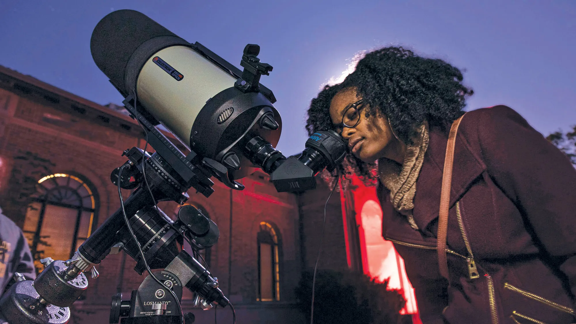 Caprice Phillips, a young black woman wearing her hair curly, a fall coat and glasses, peers into the scope of a telescope. Behind her the sky appears purple