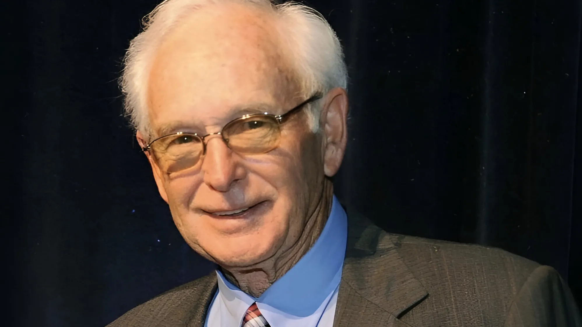 Wearing a suit, Gene Millard smiles at the person taking the photo. He's an older white man with white hair and glasses. He looks like he'd be a fun grandpa.