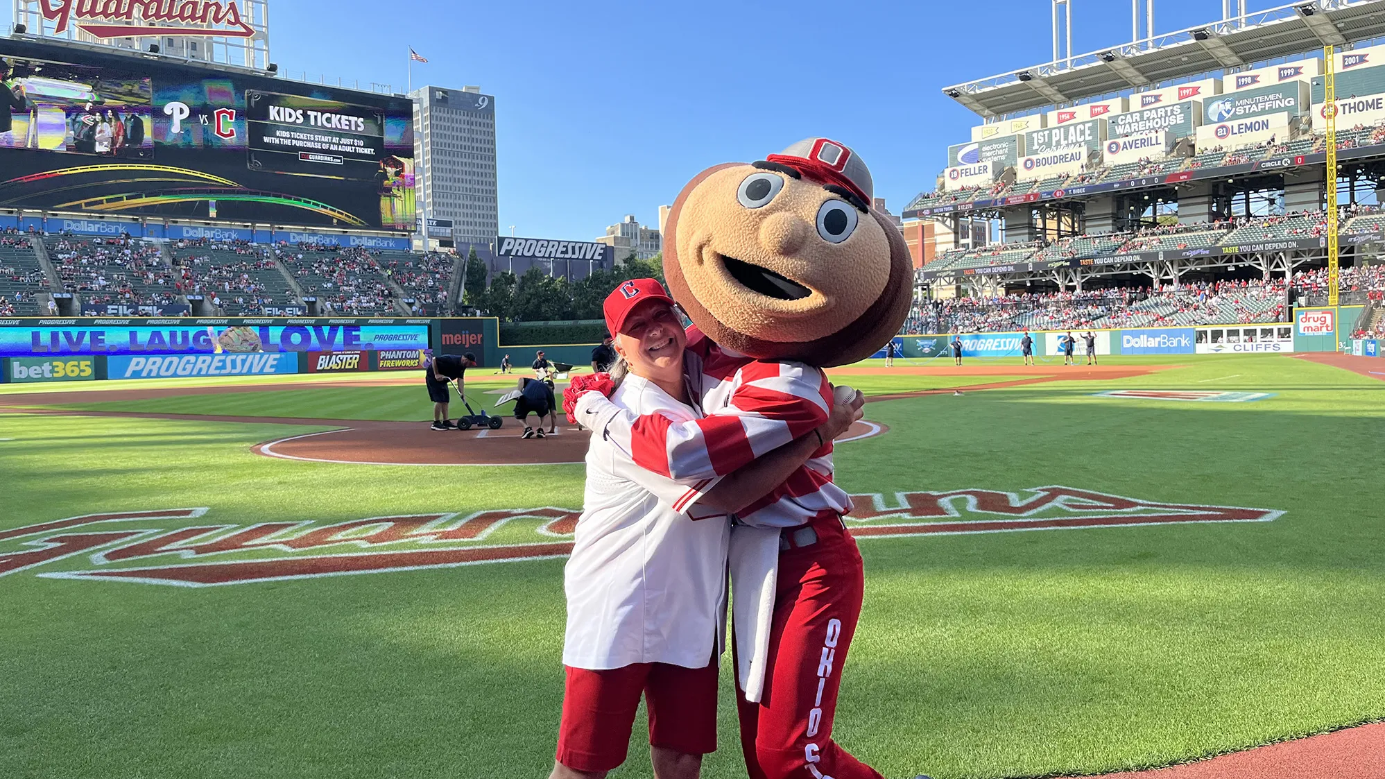 Molly Ranz Calhoun, wearing a baseball cap, T-shirt and shorts, laughs as she gives Brutus Buckeye, Ohio State's mascot with a buckeye-shaped giant head, a hug at the Cleveland Guardians' baseball stadium. Molly is a middle-age white woman with shoulder-length hair and a friendly smile.
