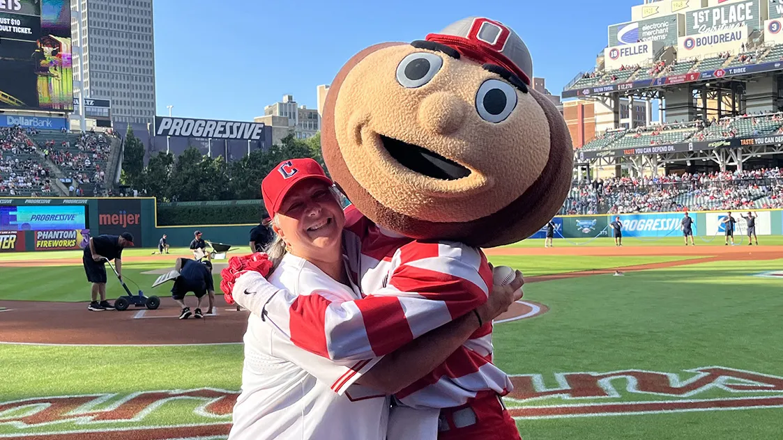 Molly Ranz Calhoun, wearing a baseball cap, T-shirt and shorts, laughs as she gives Brutus Buckeye, Ohio State's mascot with a buckeye-shaped giant head, a hug at the Cleveland Guardians' baseball stadium. Molly is a middle-age white woman with shoulder-length hair and a friendly smile.