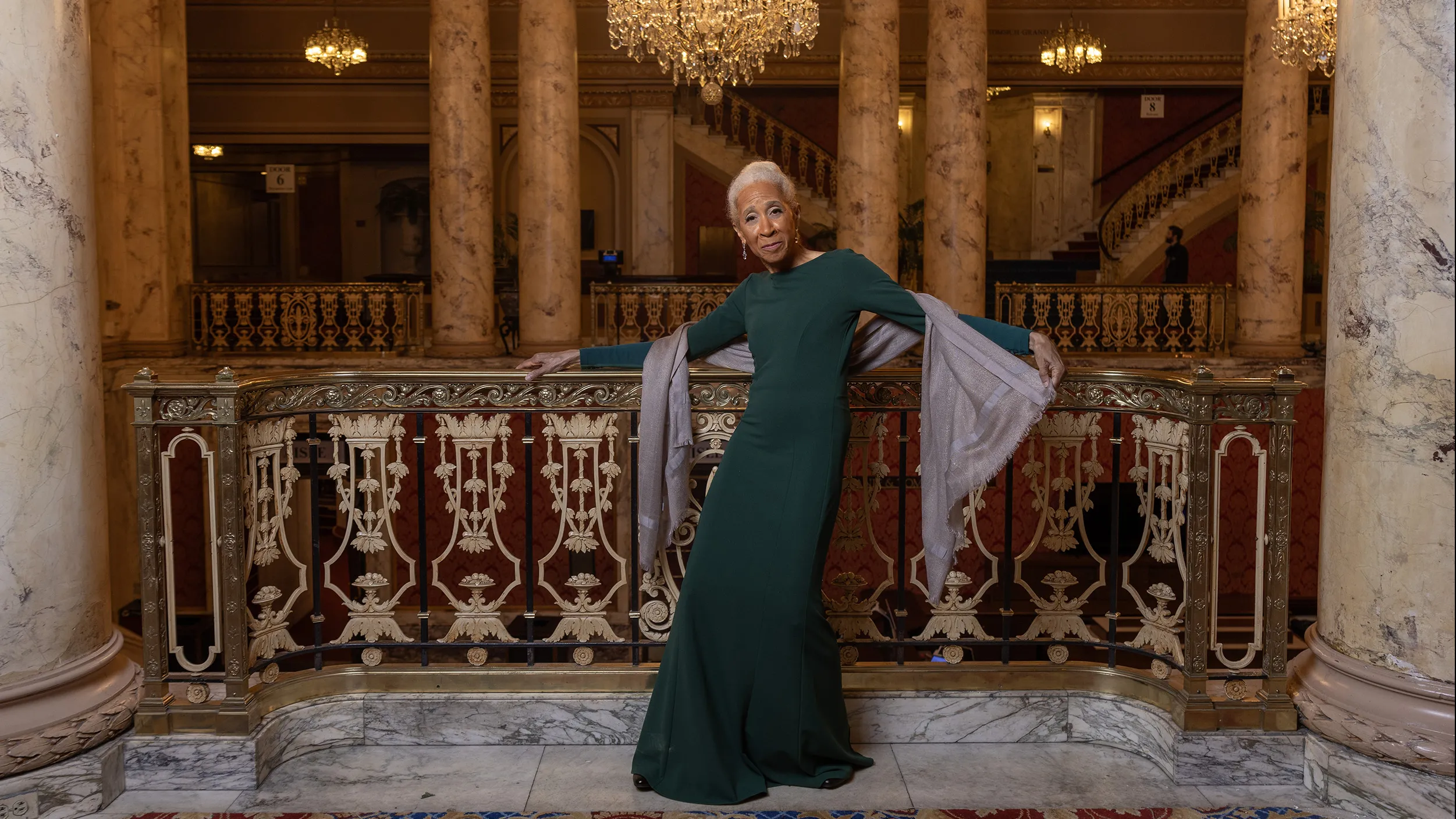 Dianne McIntyre, a slender and stylish black woman, poses along an ornate railing in a historical theatre with big marble columns and intricate detailing. Dianne is an attractive older woman. She has white hair pulled back in a bun, long legs and her body is curved as if in motion, with her arms outstretched. She wears a sleek column dress and one hand holds the end of a silk wrap draped around her back.