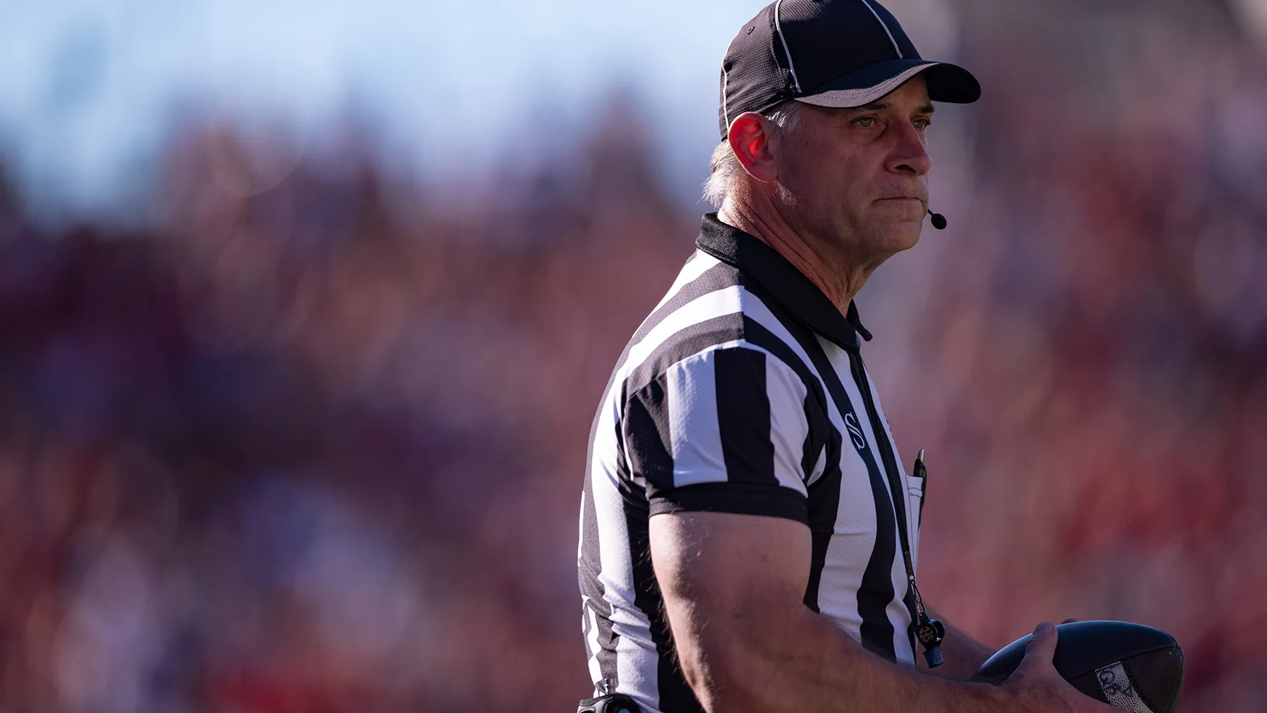 A football referee holds the ball and makes a thoughtful face as he watches something out of the photo frame. He wears a black and white striped referee’s shirt, a ballcap and a headset that leaves a small mic in front of his mouth to communicate calls. He's a white man with white hair and muscular arms.