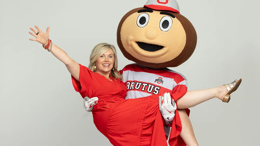 Tracy Stuck, a blond-haired white woman with a big smile and a scarlet dress, laughs as Brutus Buckeye hoists her in a cradle hold.