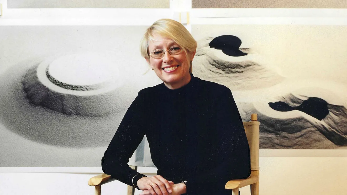 Connie Zehr, a white woman with short blond hair, oval glasses and a big smile, sits on a director's-style folding chair in front of photos of her works. They are all sand and they are mounded in different formations. They could be ancient or alien structures, or foot prints into mounded sand--they're abstract and so open to interpretation.