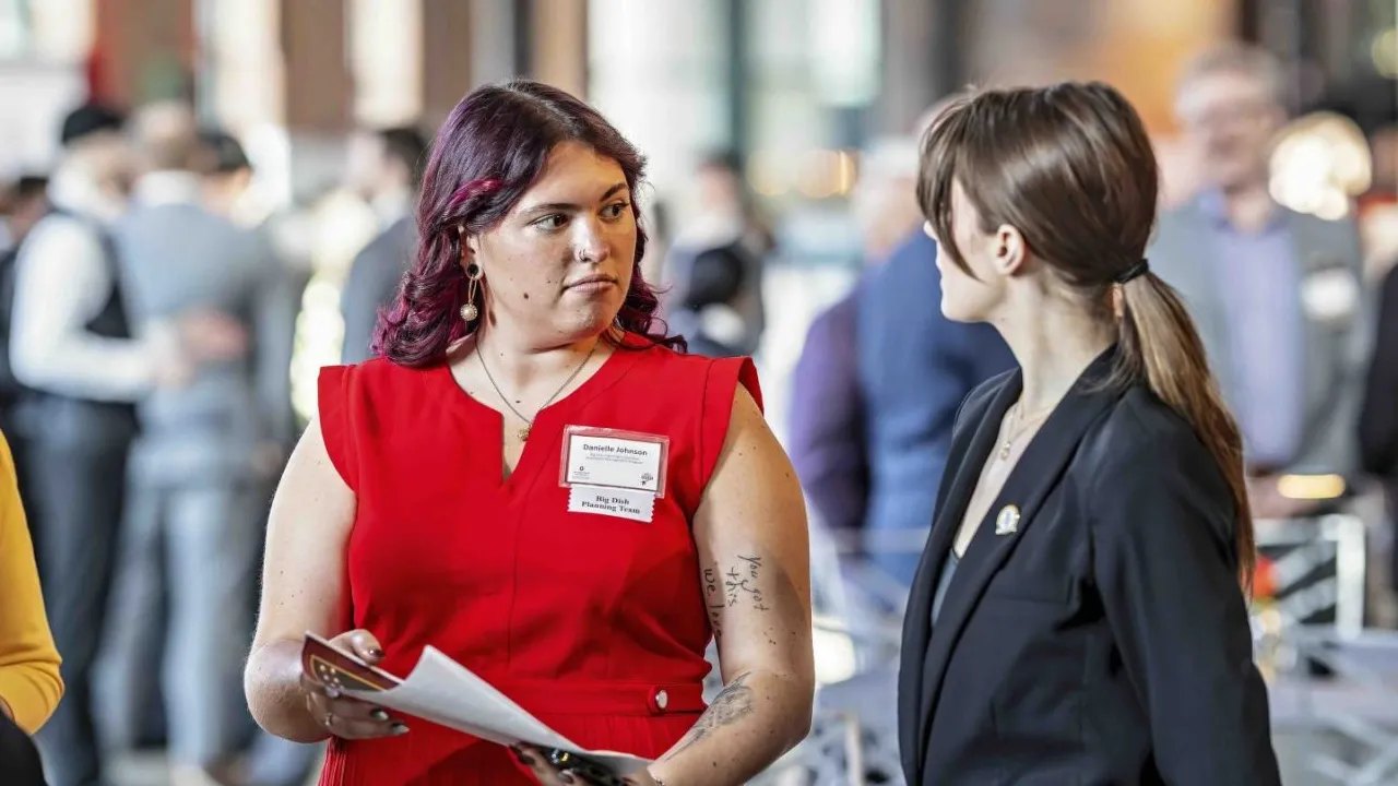 Danielle Johnson wears a scarlet dress and a focused expression as she talks to another student leader at an Ohio State hospitality event they coordinated. People dressed in suits can be seen in the background.