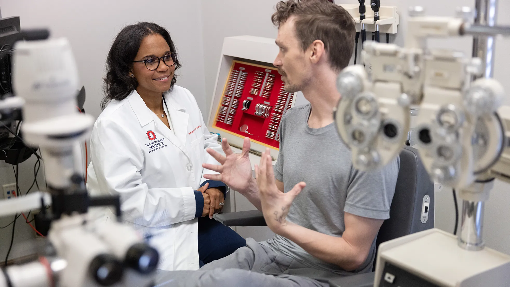 Dr. Heather Bowen-Jones smiles as she makes eye contact with the man talking with her. They both sit inside an optometry exam room. He's a white man wearing a T-shirt and gesturing with both hands. She's a Black woman wearing glasses and a doctor's white coat.