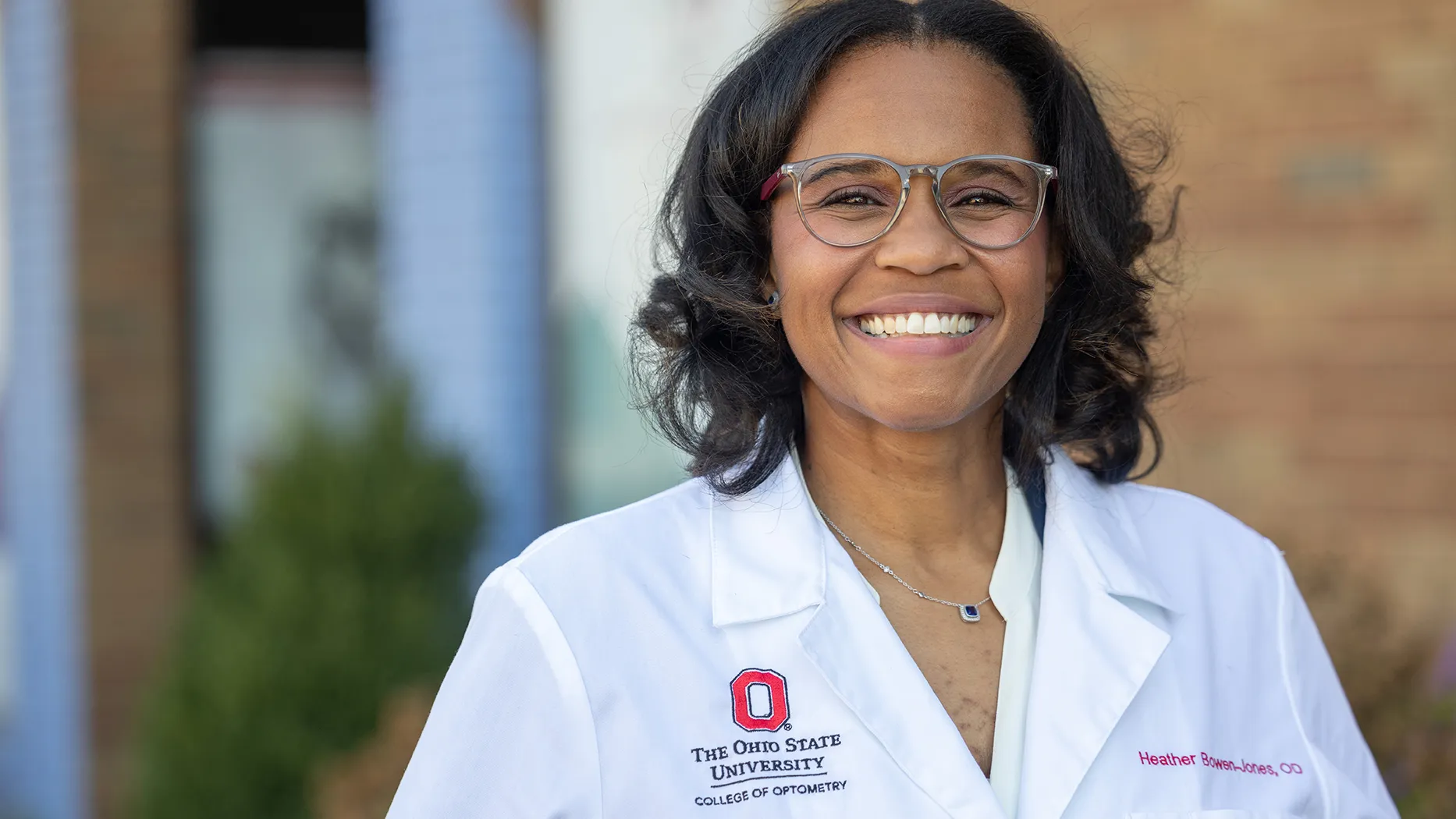 Heather Bowen-Jones ’11 OD grins for the photographer as she is photographed for a portrait. She stands in front of a medical building, wears glasses and a white doctor's coat, and she looks genuinely happy to share what she does.