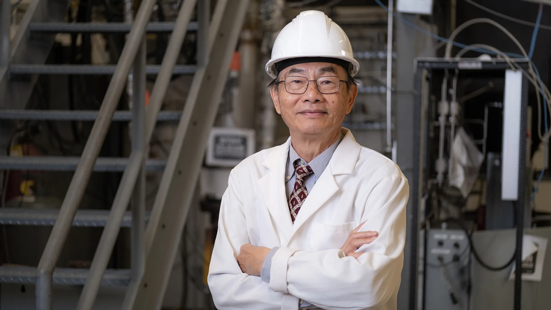 Liang-Shih Fan, an Asian man, seems quietly proud and confident, as he poses for this portrait. He wears round glasses and a white lab coat and hard hat over his suit. Behind him is the space where he works, filled with metal apparatus and tubing, as well as a staircase.