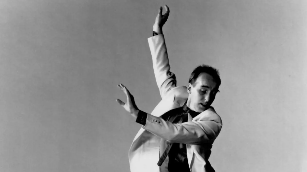 Fred Strickler tap dances with his full body slanted in a diagonal. He reaches up and out and looks down toward the stage.
