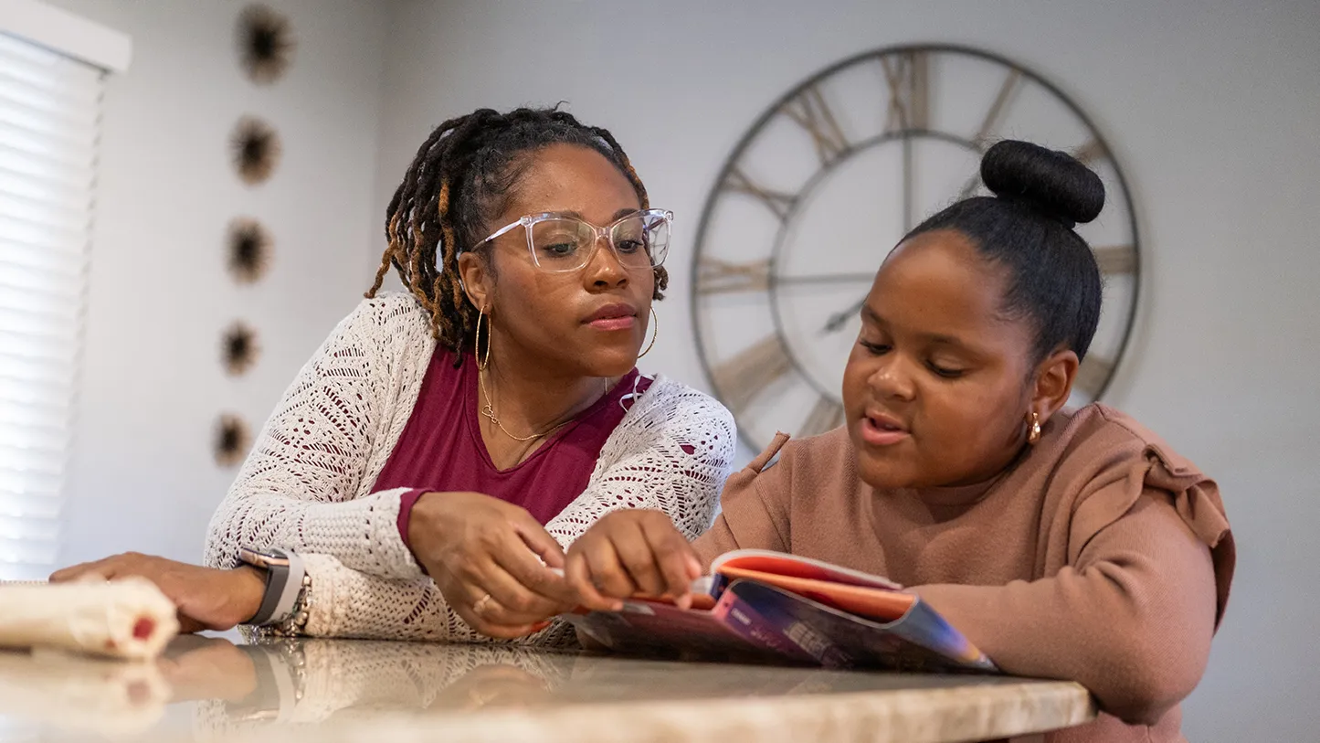 Nine-year-old Harper reads a thick children's book as her mom leans in to see the page. Harper is a cute Black girl with her hair pulled into a neat bun on top of her head, and her mom wears glasses and a pretty cardigan. They sit in their kitchen at home.