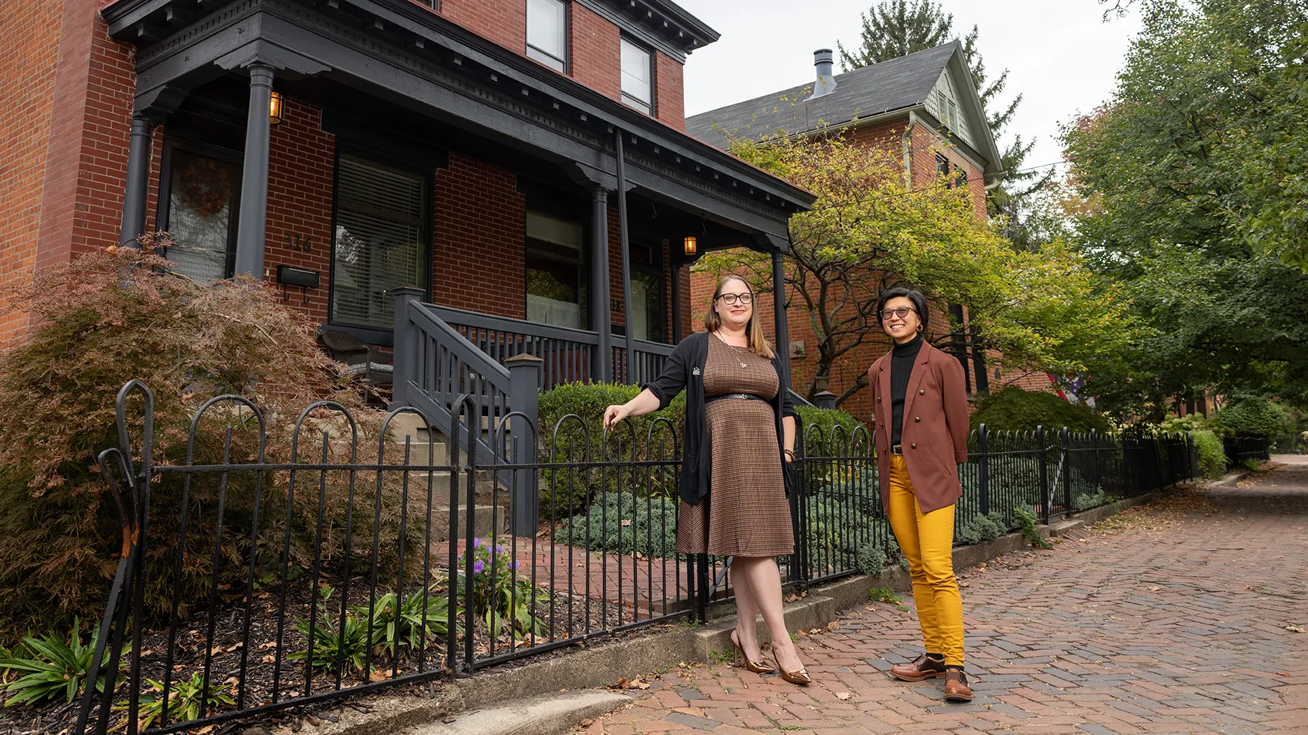 Two young people stand outside a well-kept brick duplex. They created the documentary on duplexes. One softly smiles and wears a dress, cardigan and black leather belt. The other smiles bigger and wears yellow pants and a brown suit jacket.