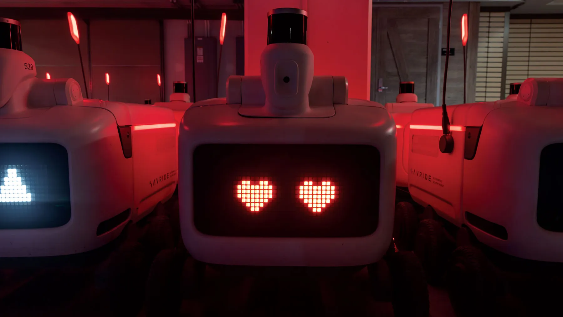 In a dark room, a plugged-in robot has pink hearts for eyes on its face screen. Red light shines around it.