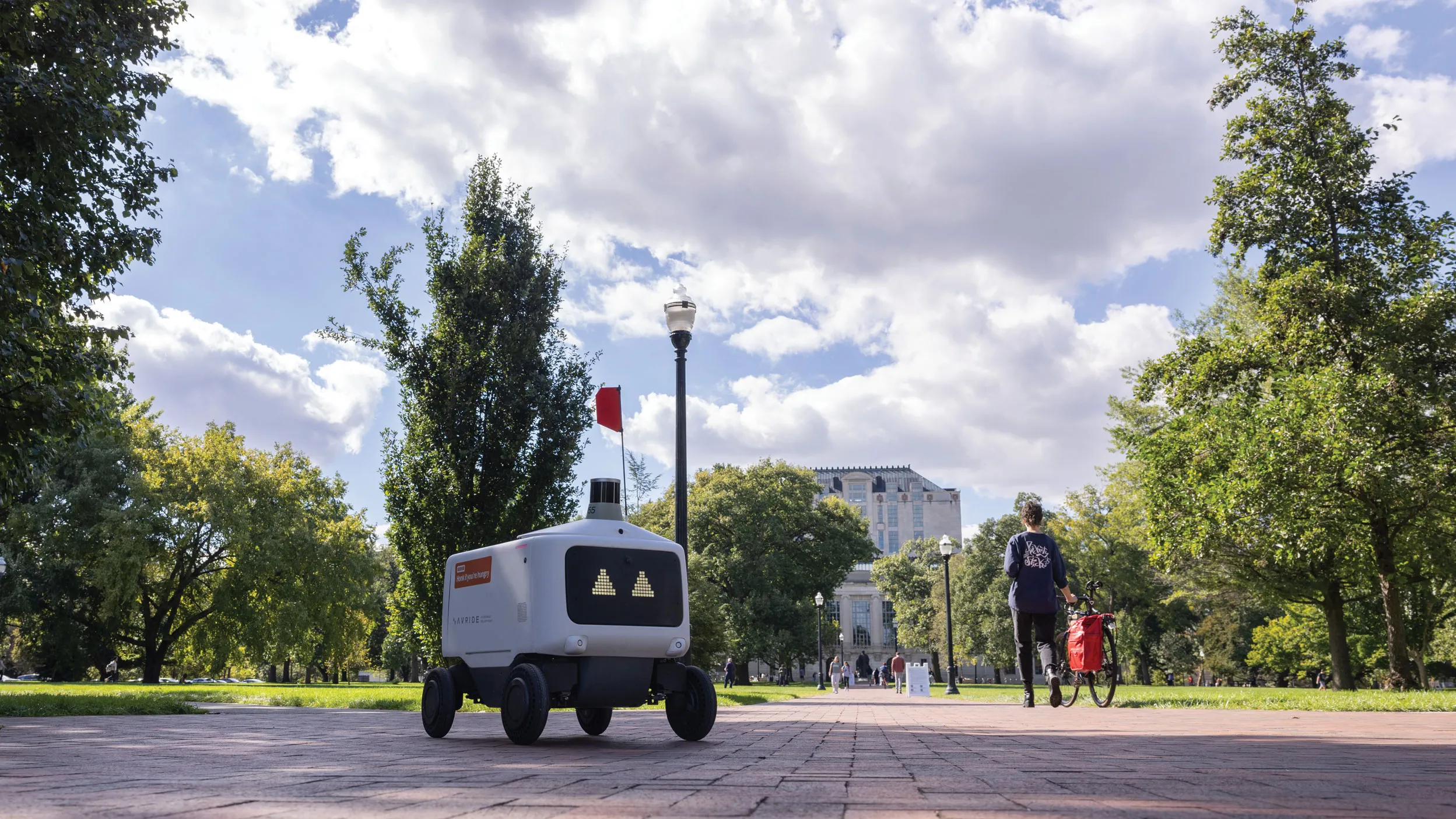 A lone food rover, which is a plastic-covered rectangle with a screen face showing trinagle-shape eyes, makes its way across the oval. Thompson Library and students can be seen in the distance.