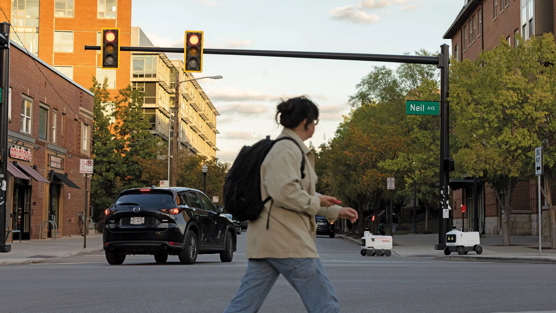Two food delivery robots venture across a street, as a vehicle turning left rolls in front of them and a student, crossing across the intersection from them, watches their progress.