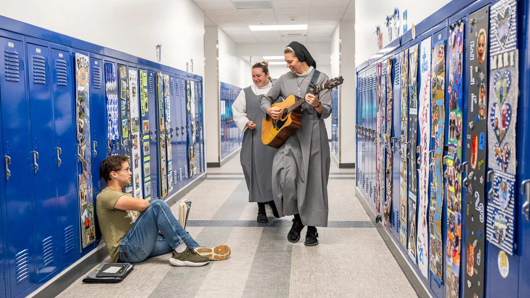 Sister and a colleague walk past lockers up the hallway. Sister plays a guitar. A high school teen sits on a floor in front of an open locker.