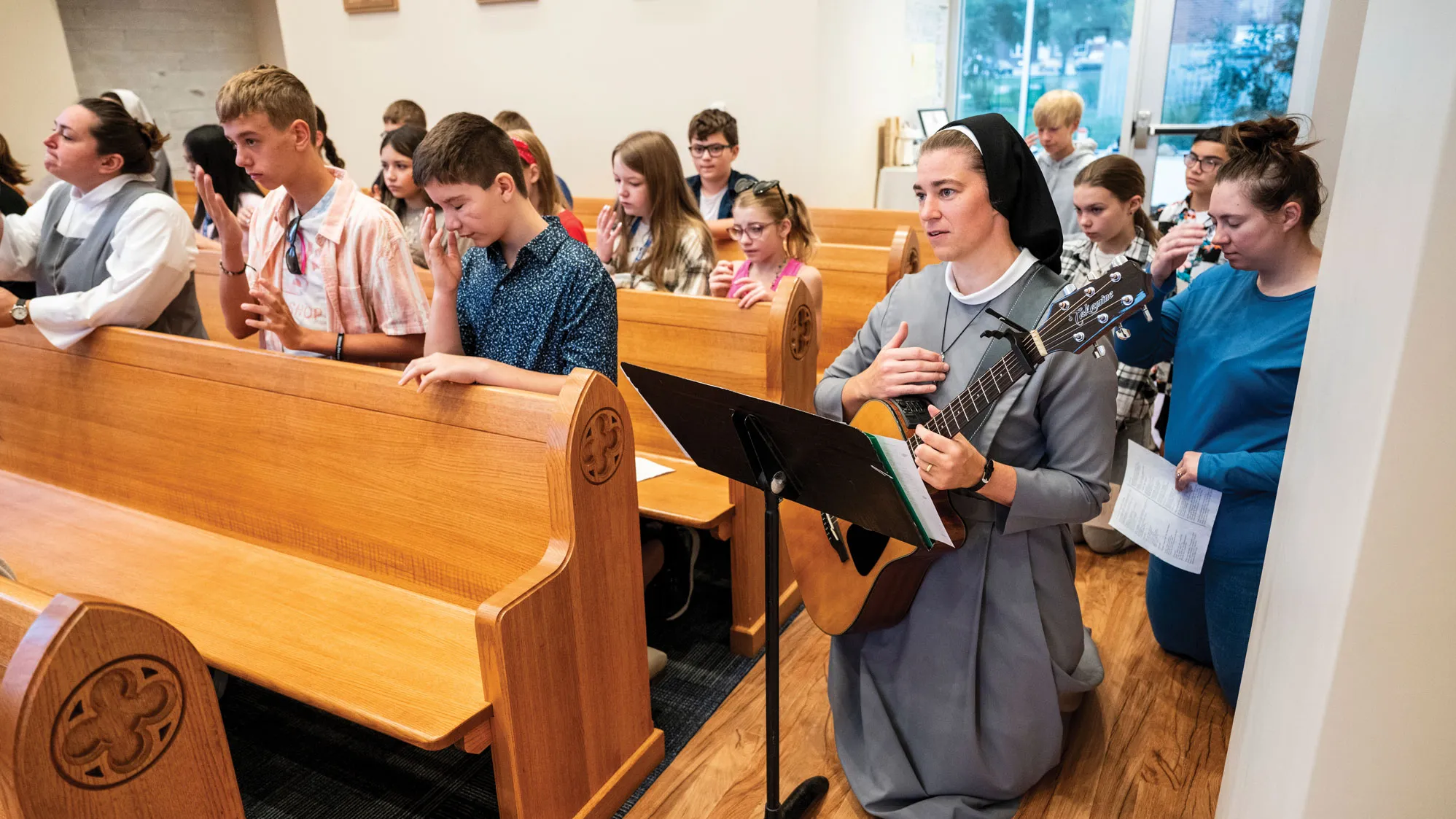 Sister kneels in her habit on the hard wood of a chapel floor. She's making the sign of the cross and holds a guitar. A music stand is before her. Behind her, other people kneel. In the pews next to her, teenagers kneel and make the sign of the cross. Her expression looks supremely peaceful and worshipful.