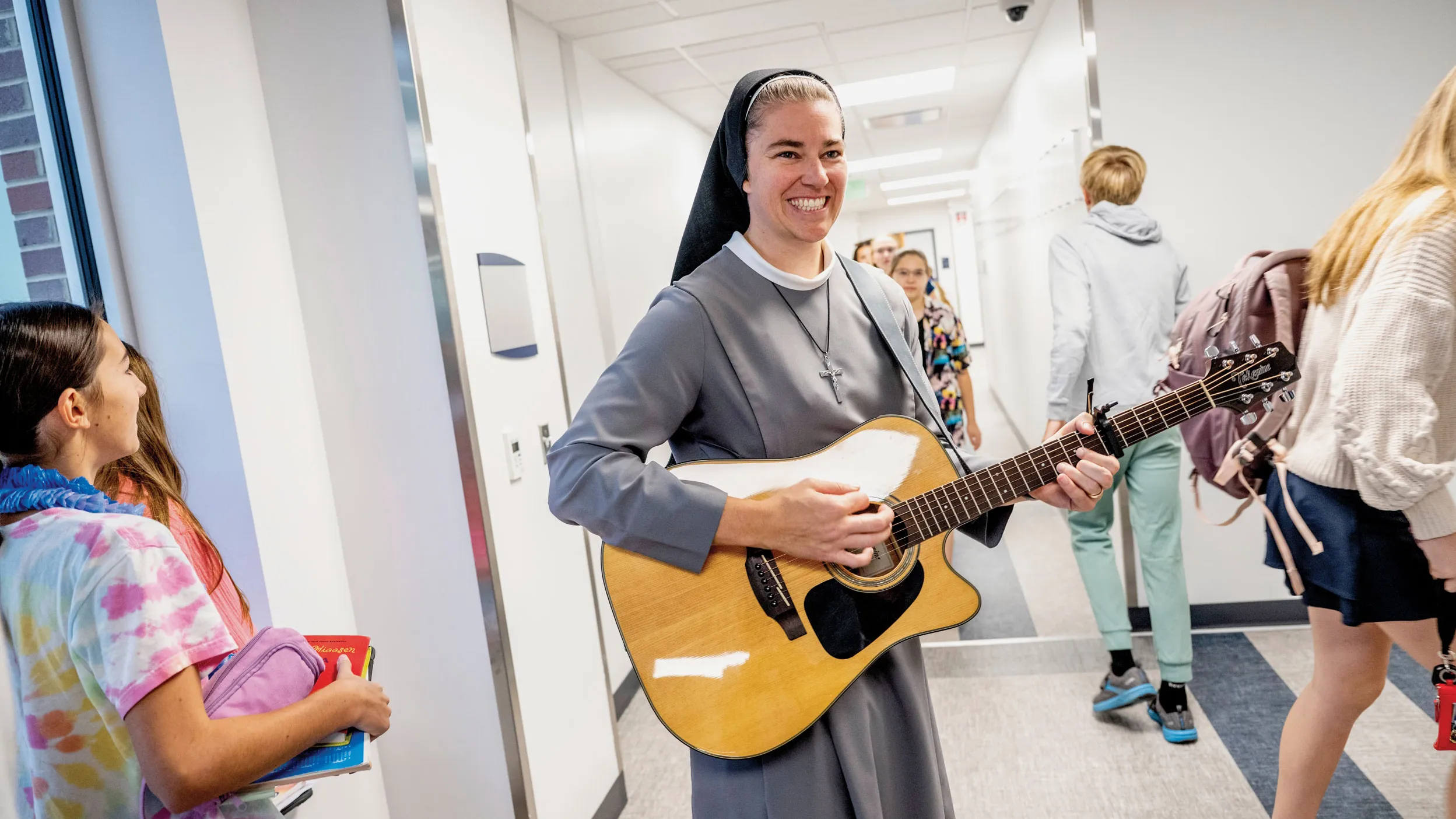 Sister Mary Xavier Schulze is a white woman wearing a veil and habit in shades of gray, white and black. She smiles big and plays a guitar as she walks the halls of the school she works in, as kids rush past her.