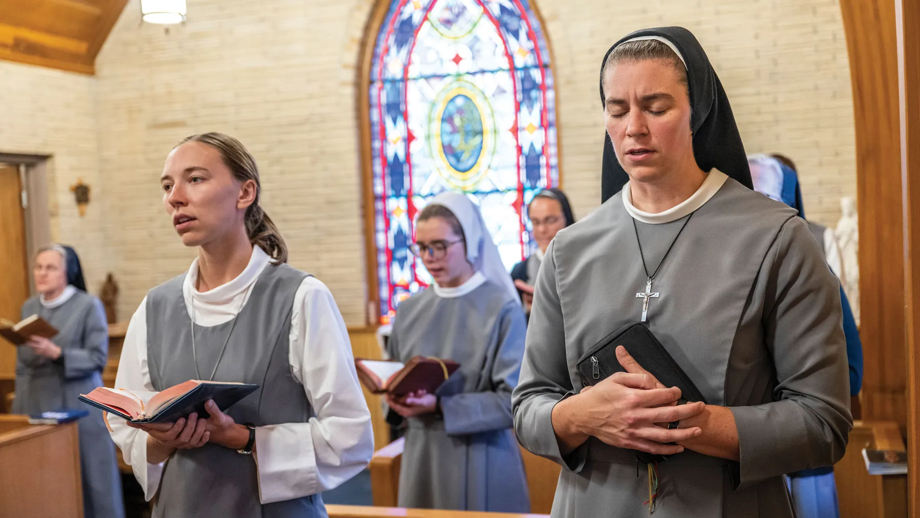 Sister prays fervently at a prayer service with other sisters. A stained glass window behind them shines light in on them.